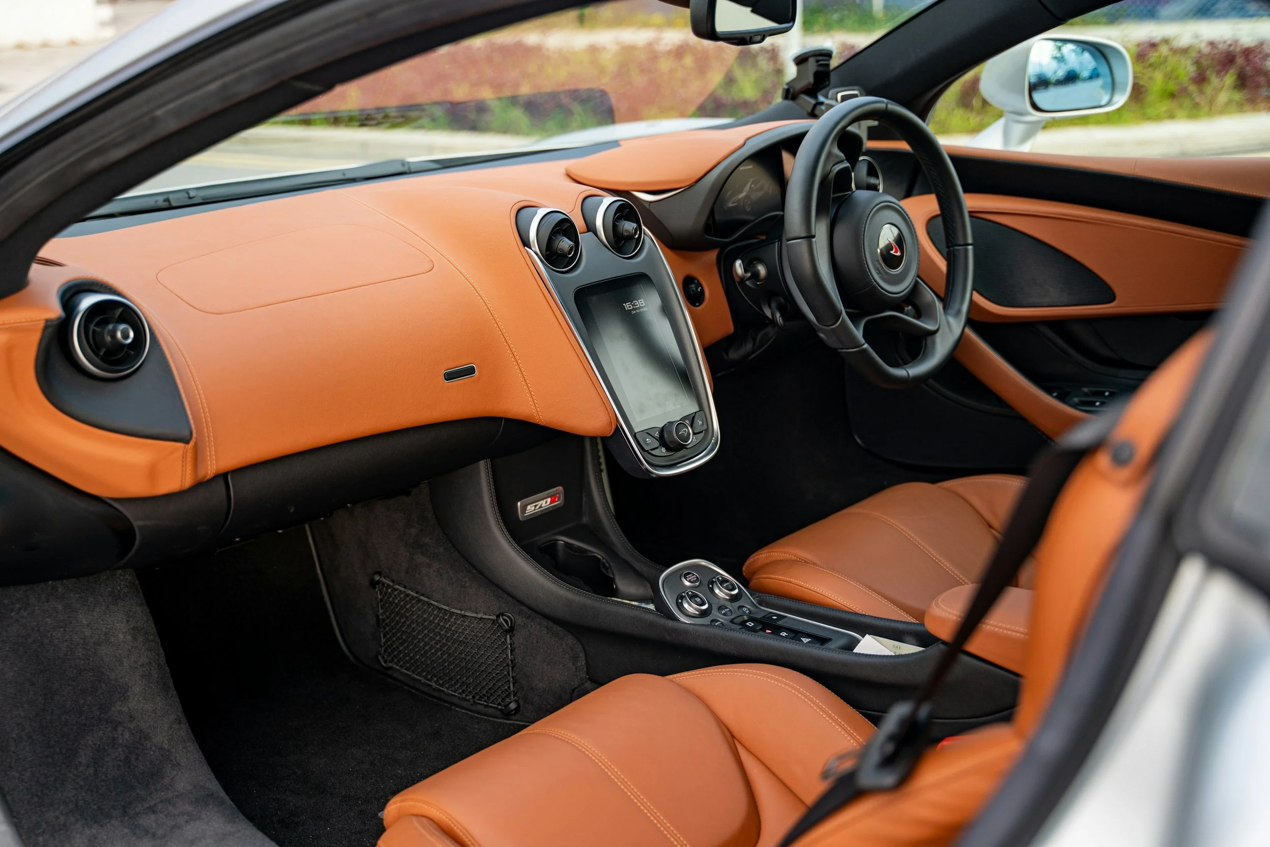 Interior of a sports car with tan leather seats, a central touchscreen display, and a black steering wheel with a logo in the center.