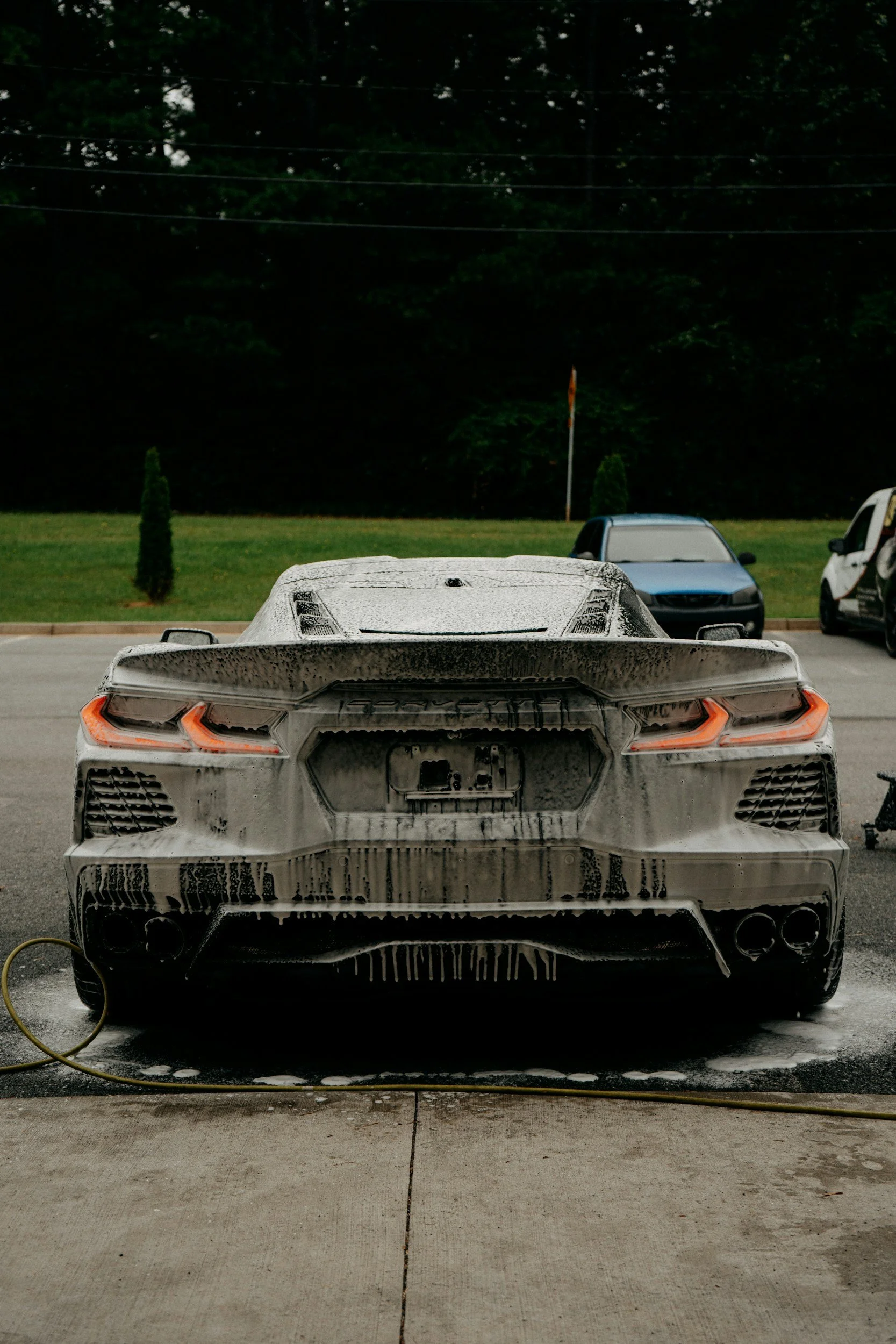 A white sports car covered in soap suds during a car wash. The car is parked on a driveway with a grassy area and trees in the background.