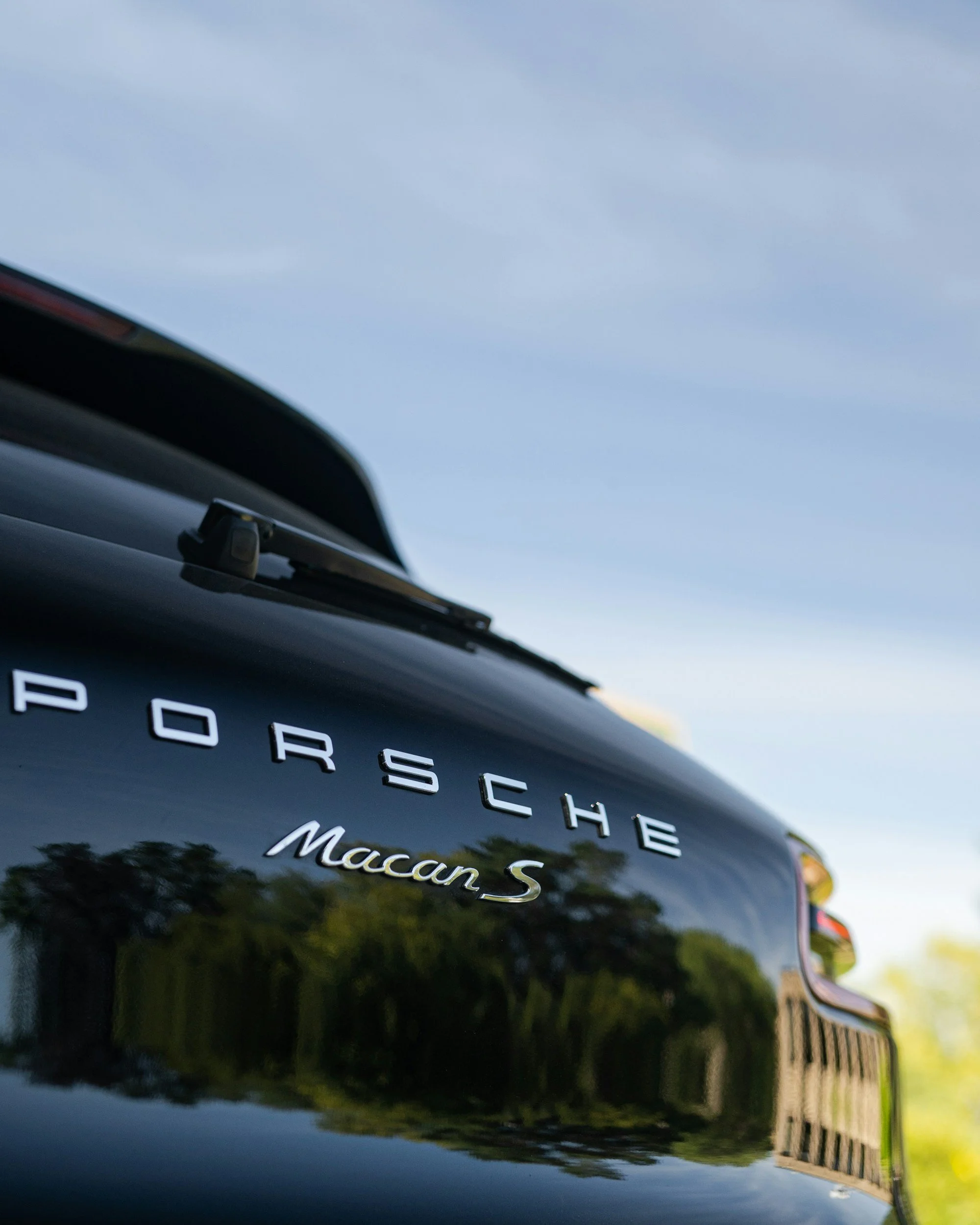 Close-up of the rear of a black Porsche Macan S SUV, with the Porsche logo and model name reflected on the shiny surface.