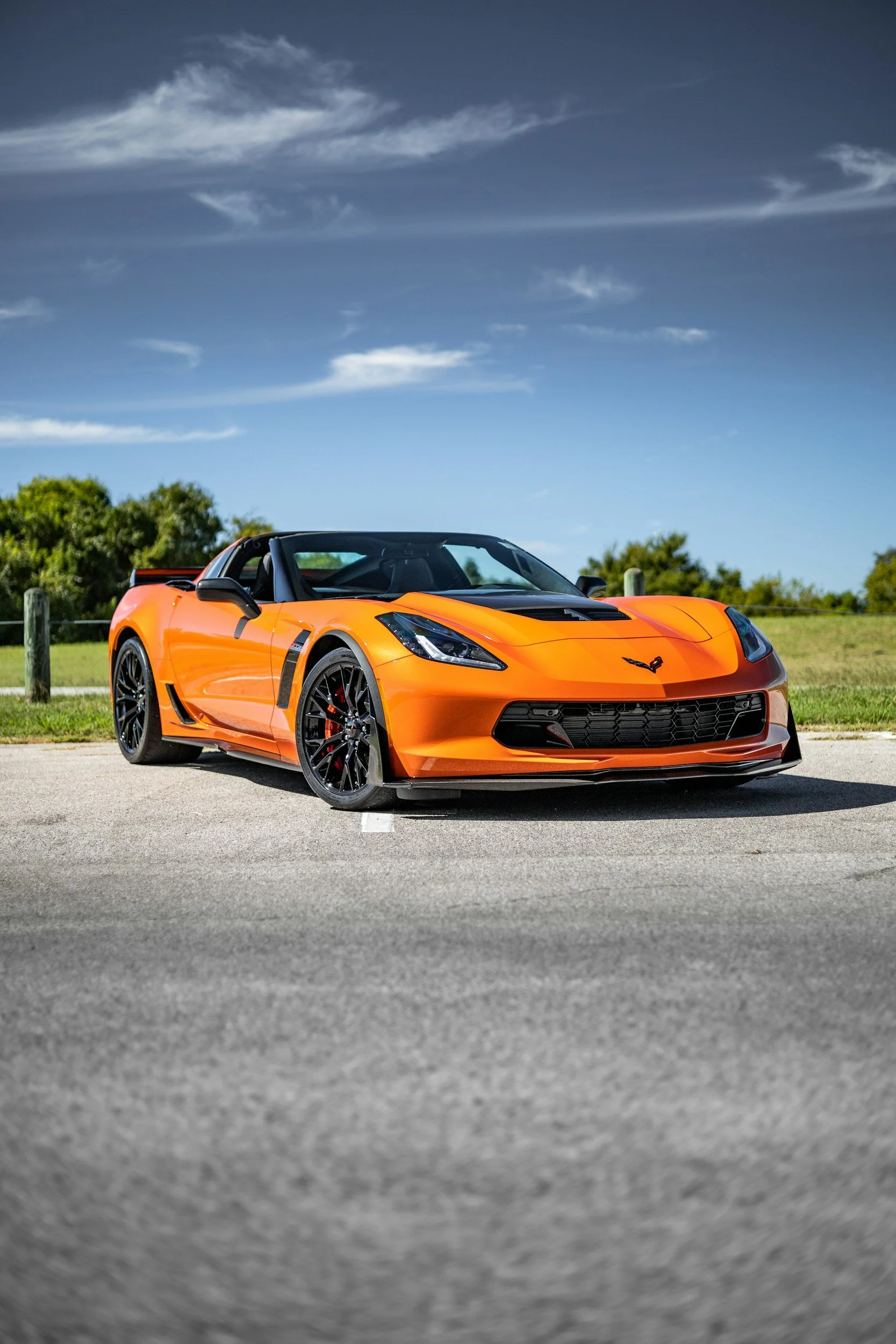 An orange Chevrolet Corvette sports car parked on a paved surface with grass and trees in the background under a partly cloudy sky.
