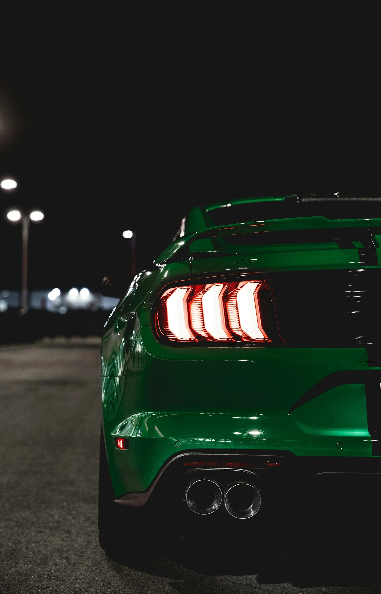Nighttime image of the rear of a green sports car with distinctive taillights and dual exhaust pipes, parked outdoors.