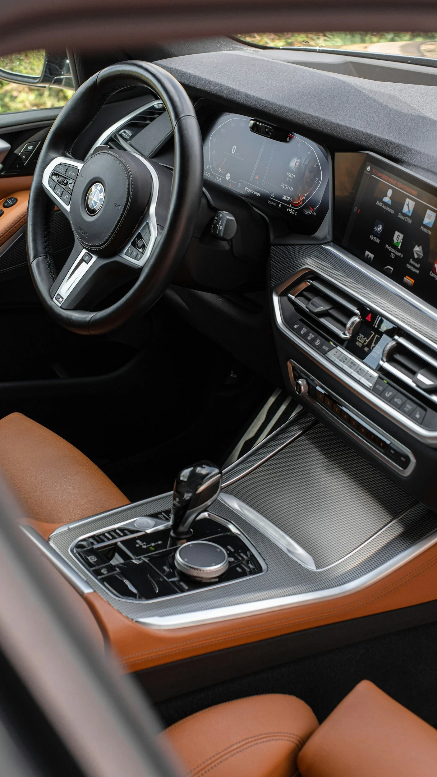 Interior view of a modern BMW car, showing the steering wheel, digital instrument cluster, infotainment screen, center console with gear shift, and tan leather seats.