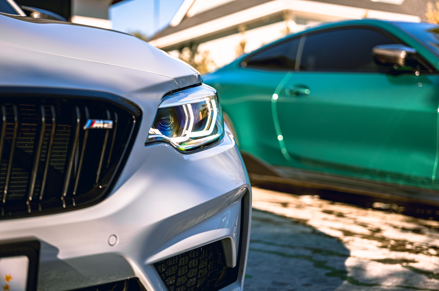 Close-up of the front of a white BMW M Series car with a black grille, and a blurred green car in the background outside on a paved surface.