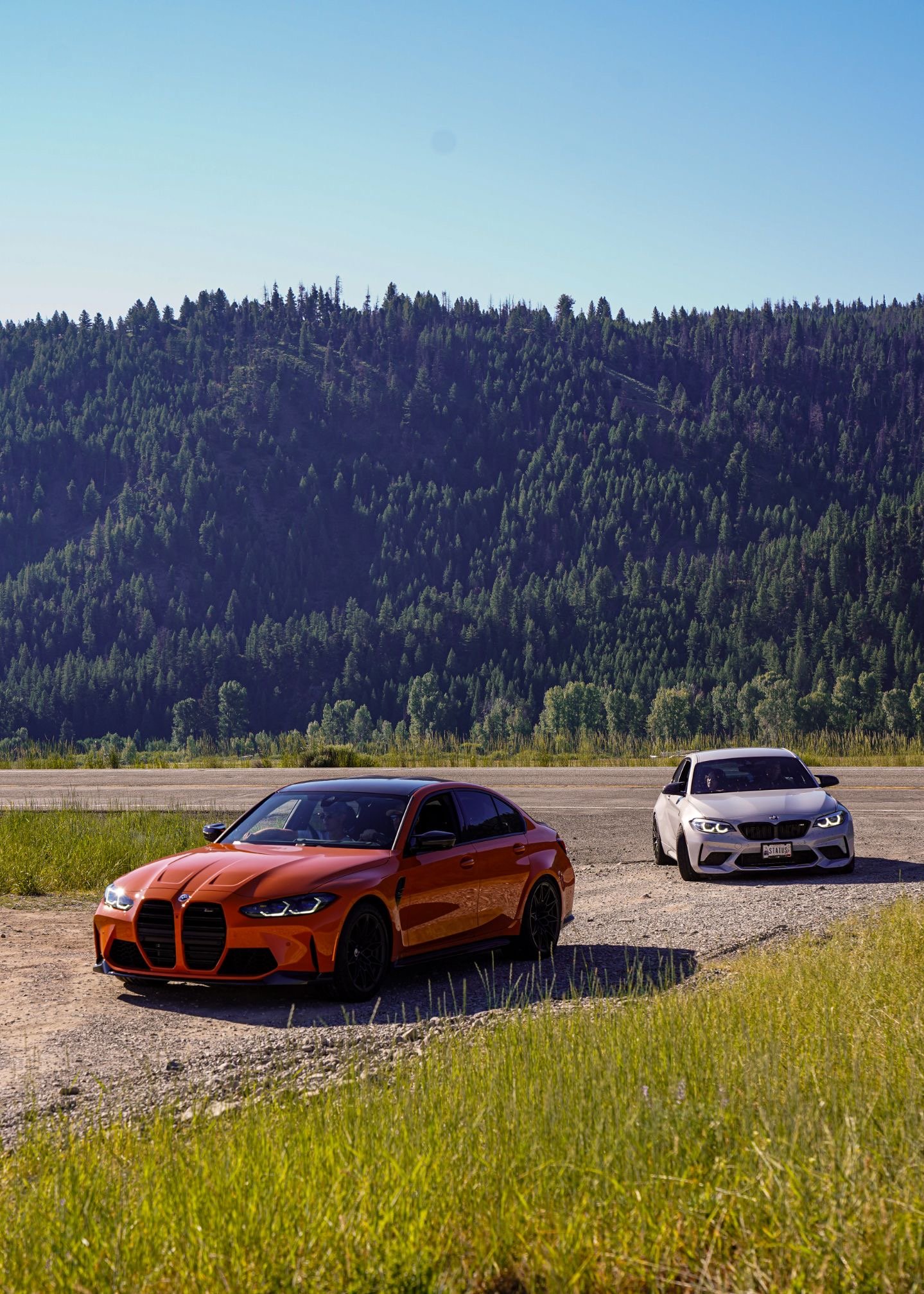 Two cars parked on a gravel area near greenery, with a mountain covered in trees in the background and a clear blue sky overhead.