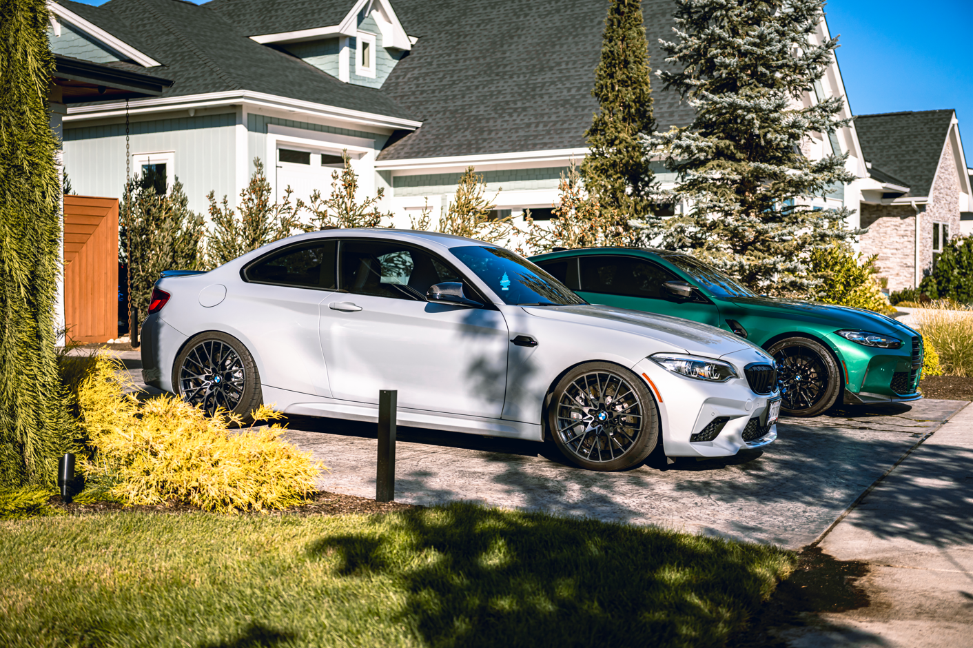 A white BMW and a green car parked in a driveway in front of a large house with a gray roof, surrounded by trees and bushes on a sunny day.