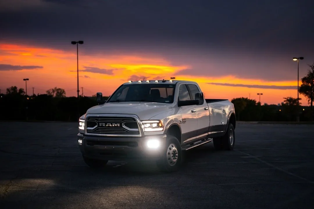 A white Ram pickup truck parked in a mostly empty parking lot during sunset with a colorful sky and some trees in the background.