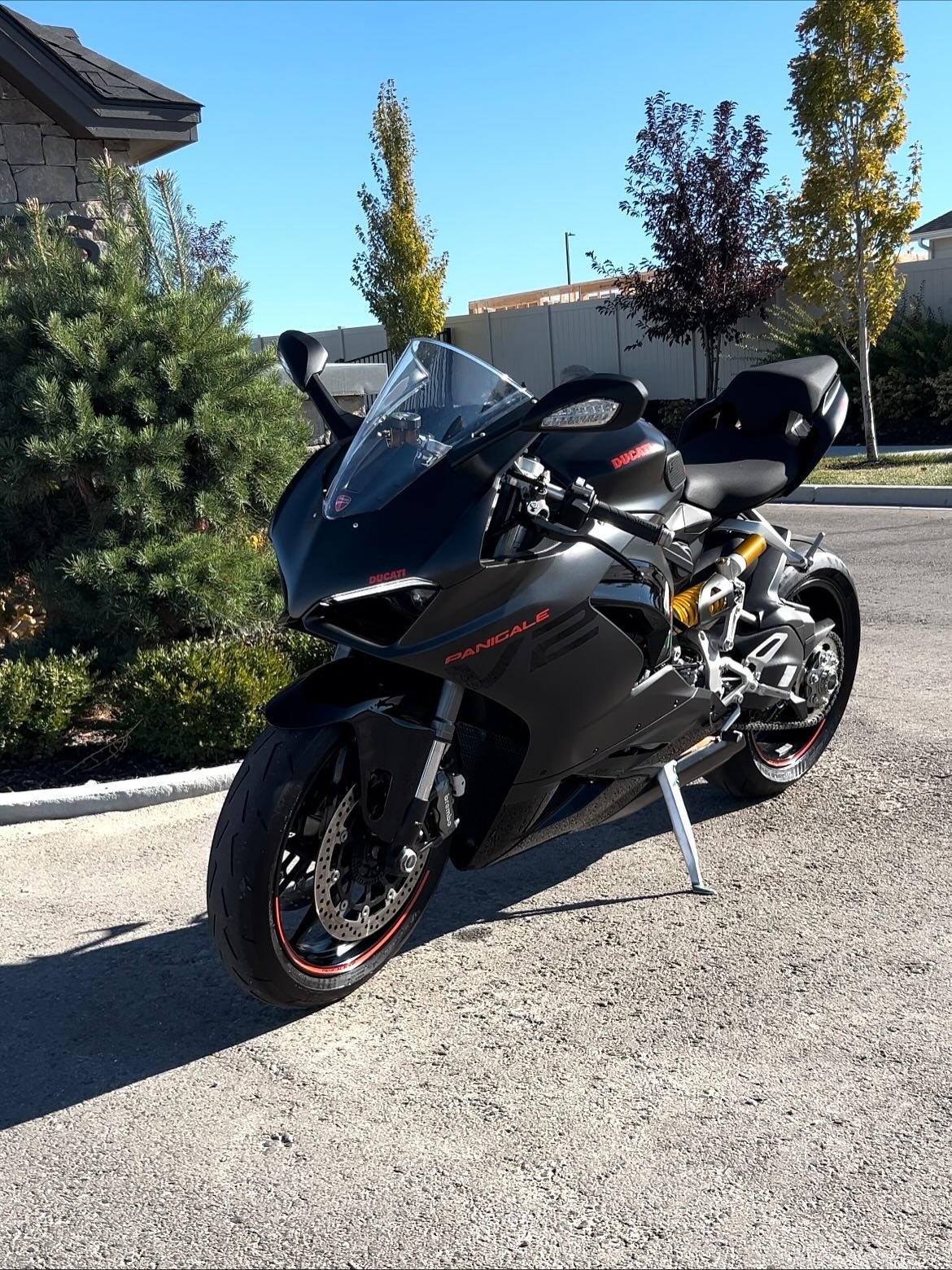 A black Ducati Panigale sport motorcycle parked on a driveway, with trees and a house in the background.