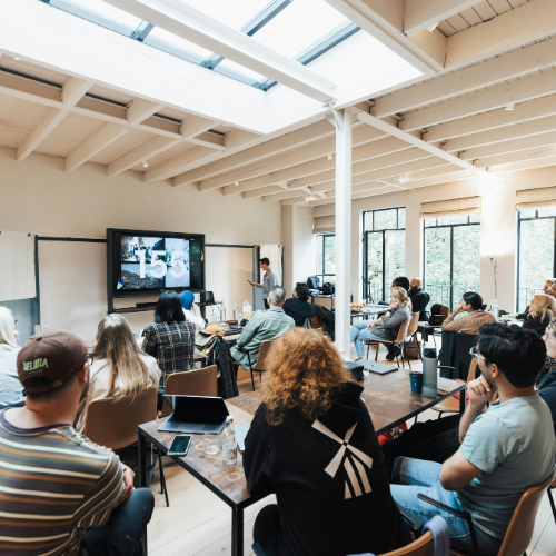 People attending a presentation in a modern, sunlit conference room with large windows and a television screen.