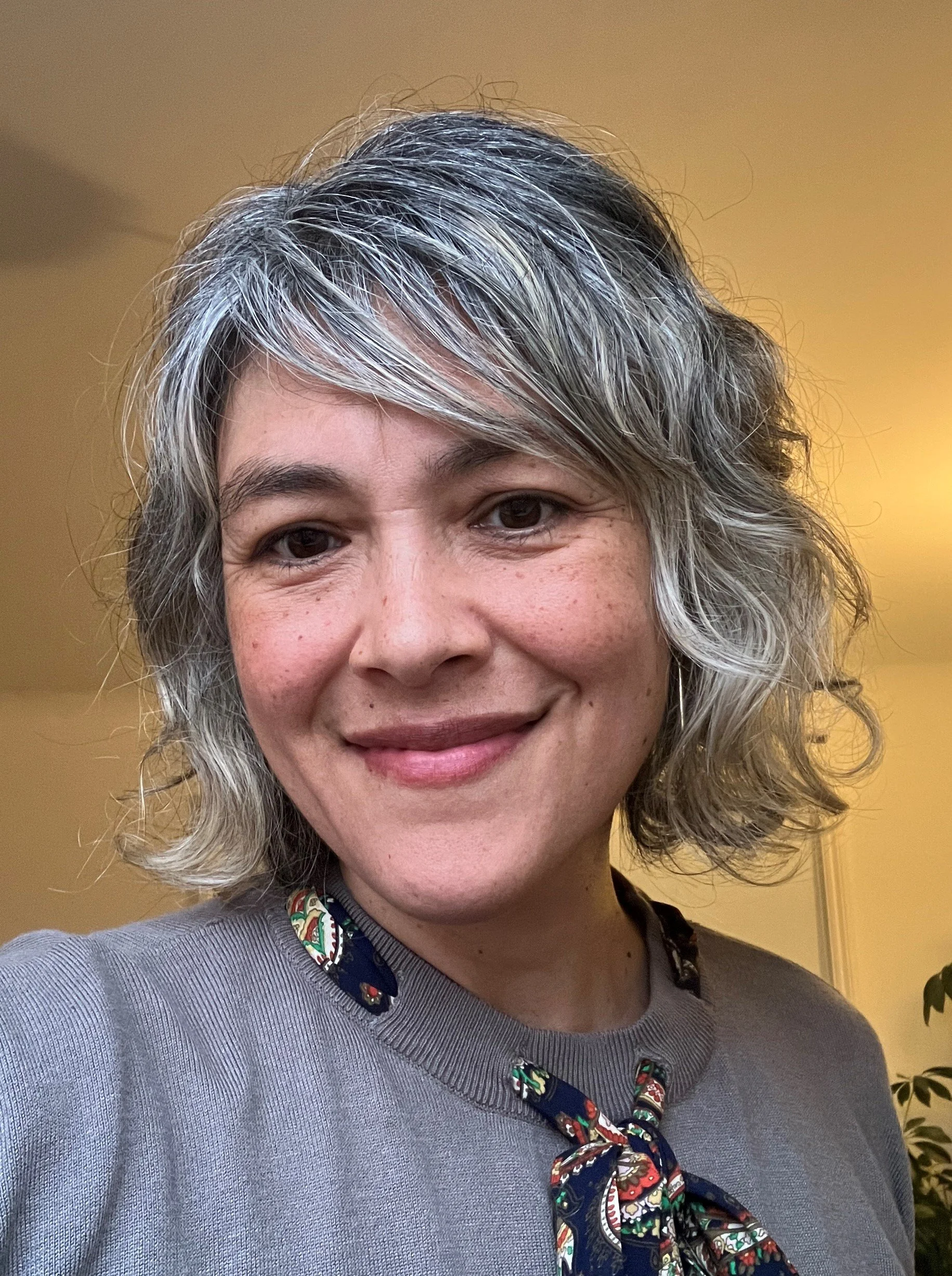 A woman with short, wavy gray hair, smiling, wearing a gray sweater with a colorful patterned scarf tied around her neck, standing indoors with a yellowish wall and a plant visible in the background.