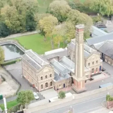 Aerial view of a historic brick building with a tall clock tower, surrounded by green trees and a parking lot.