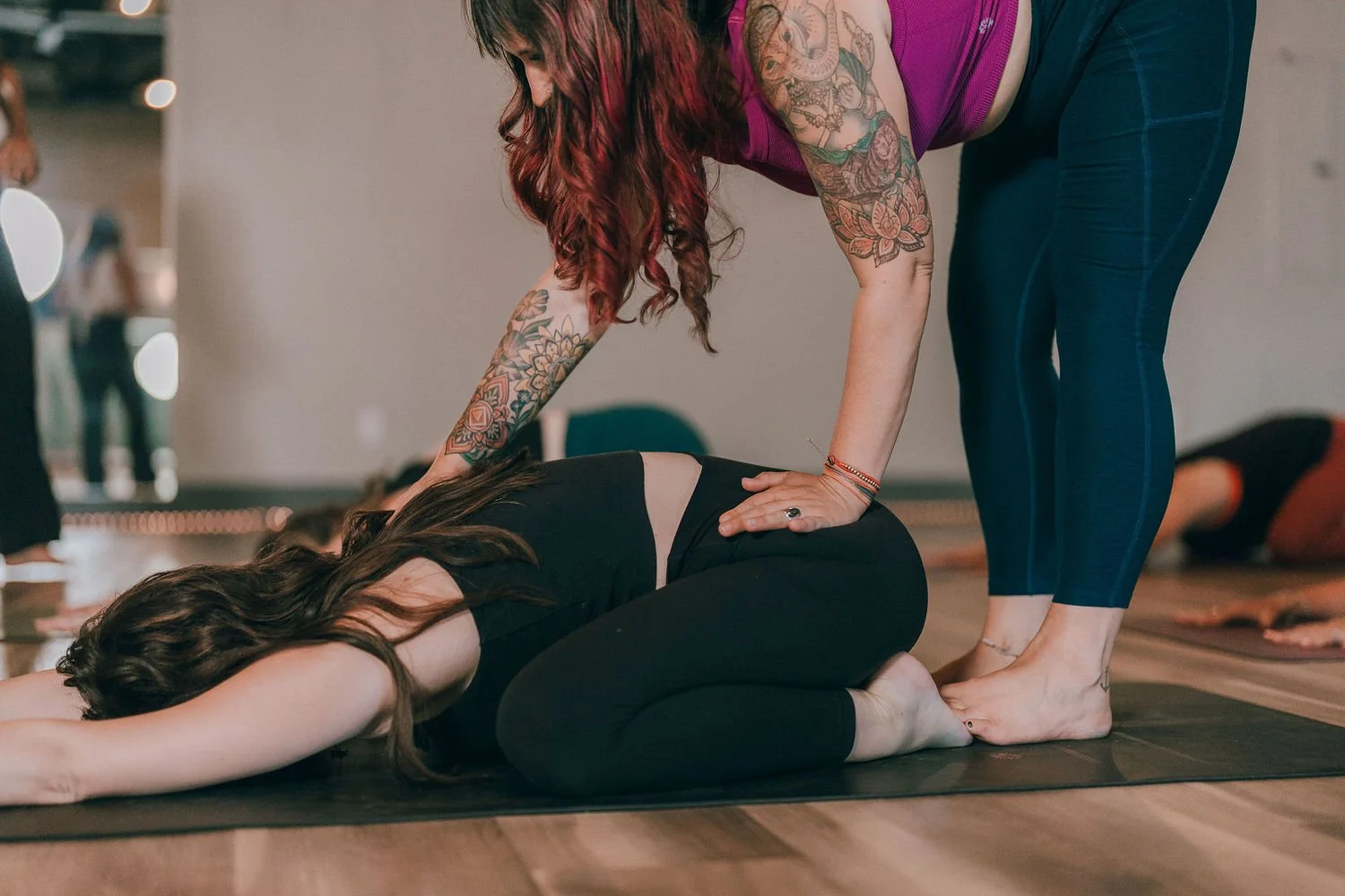 A woman with long hair lying face down on a yoga mat at Spark Hot Yoga in Snohomish WA, while another woman with tattoos assists her in a yoga pose.