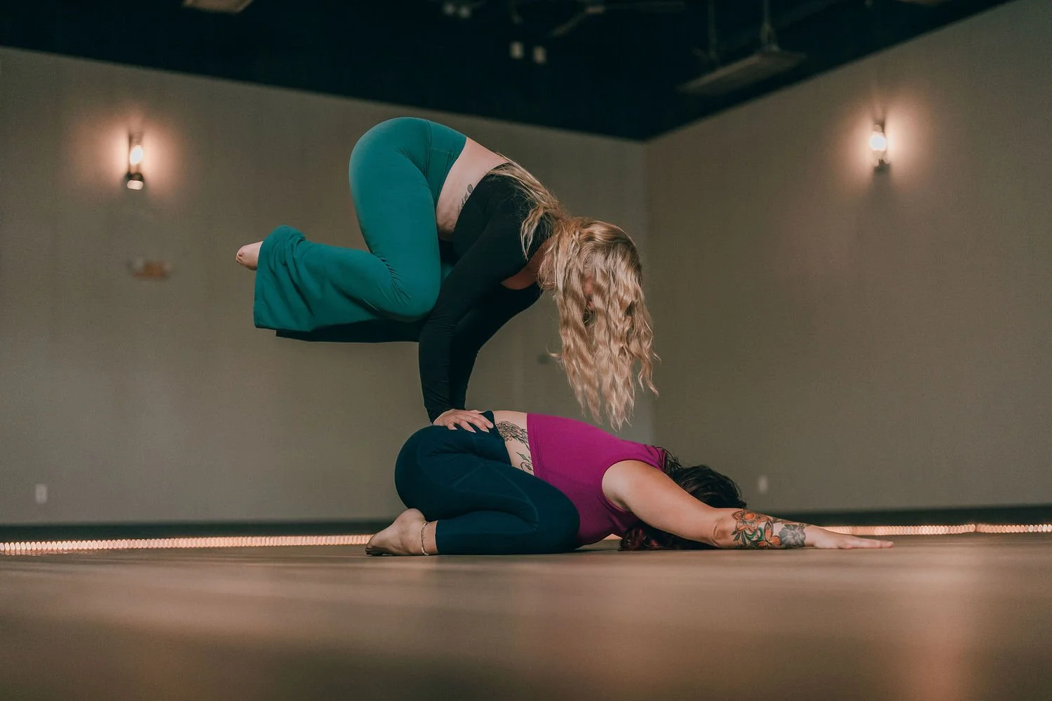 Two women practicing acro yoga indoors, with one woman on her hands and knees on the floor and the other balancing above her in a plank position, supporting her own weight.