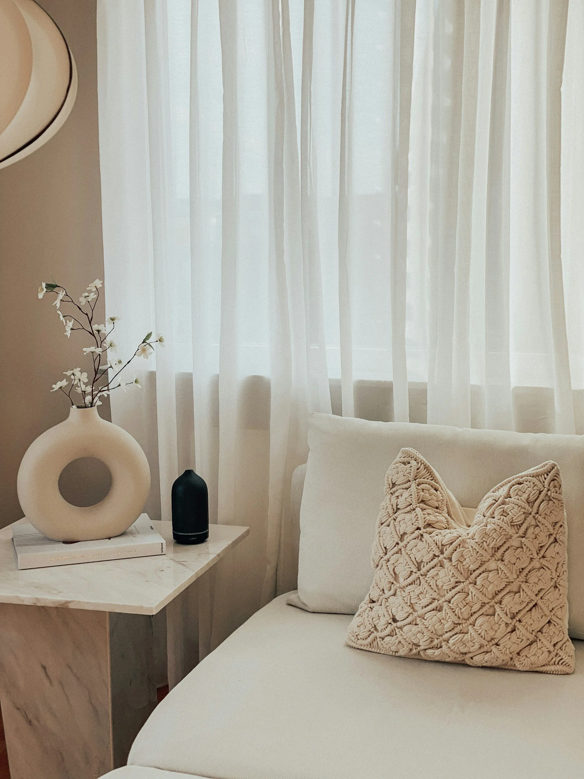 Living room corner with white curtains, a white sofa with a textured beige pillow, a small marble side table holding a vase with white flowers, a black diffuser, and a book.