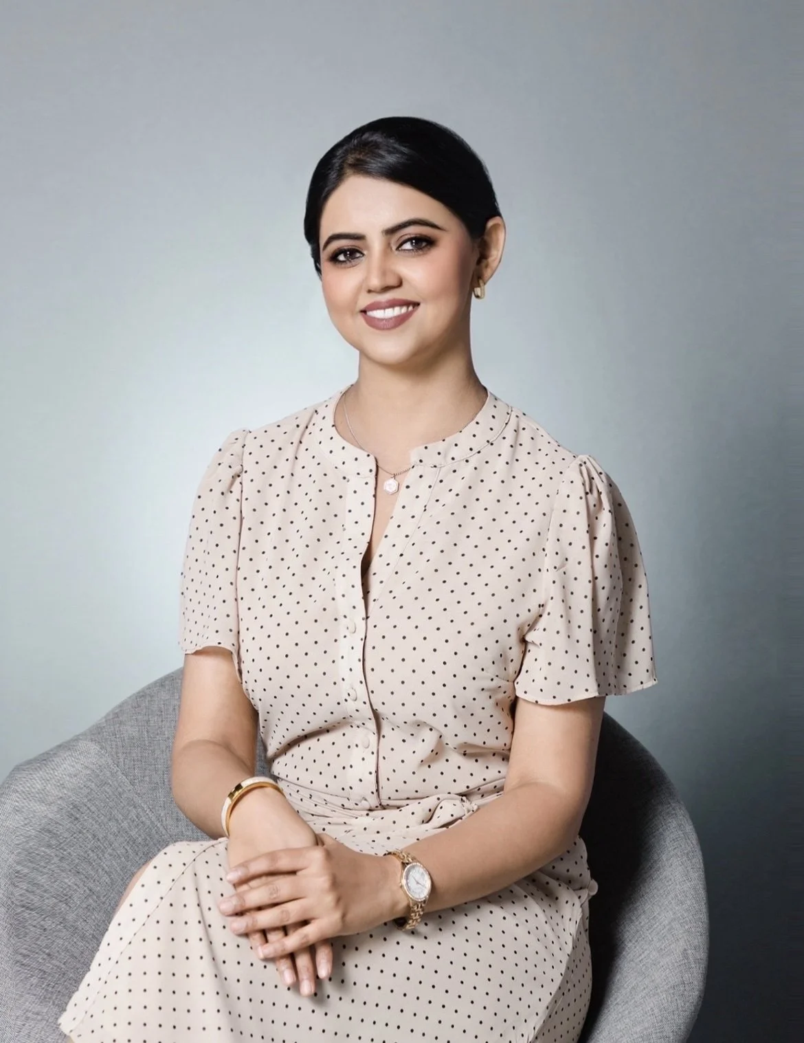 A woman with short black hair, wearing a beige polka-dot dress, seated on a gray chair, smiling with a plain gray background.