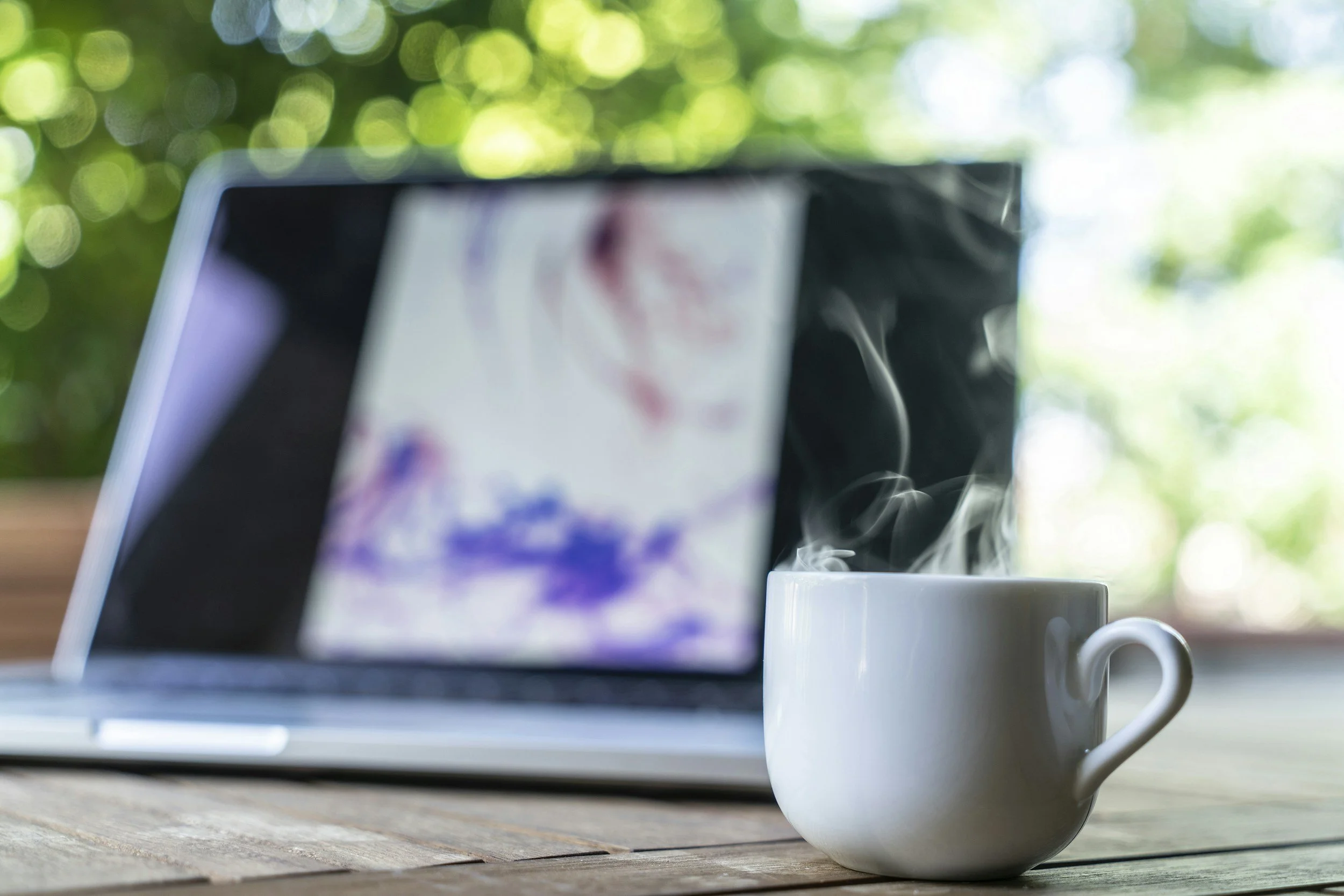 A steaming white coffee mug placed on a wooden table with a laptop in the background outside with greenery and sunlight.