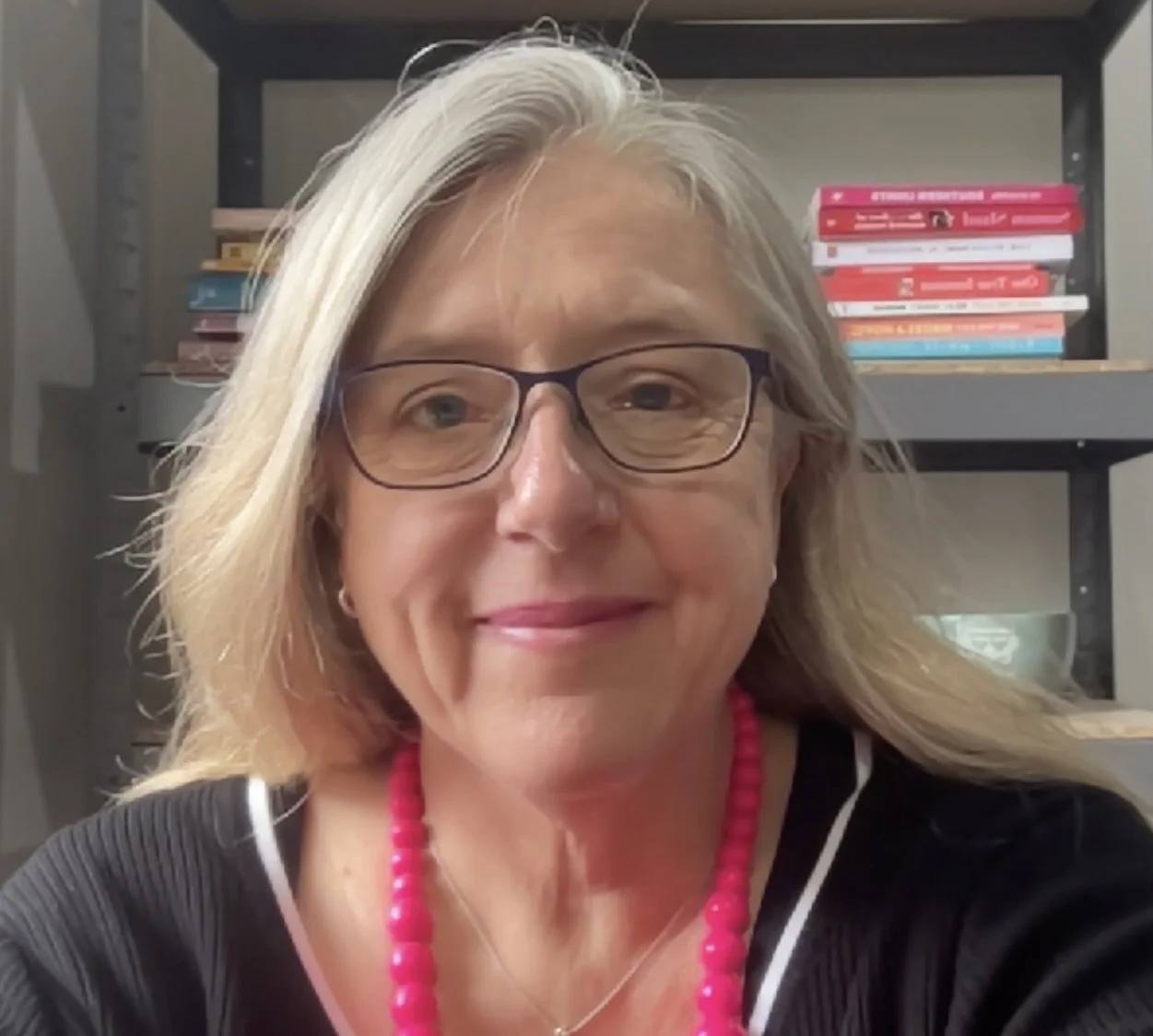 A woman with glasses and blonde hair smiling in front of a bookshelf with colorful books
