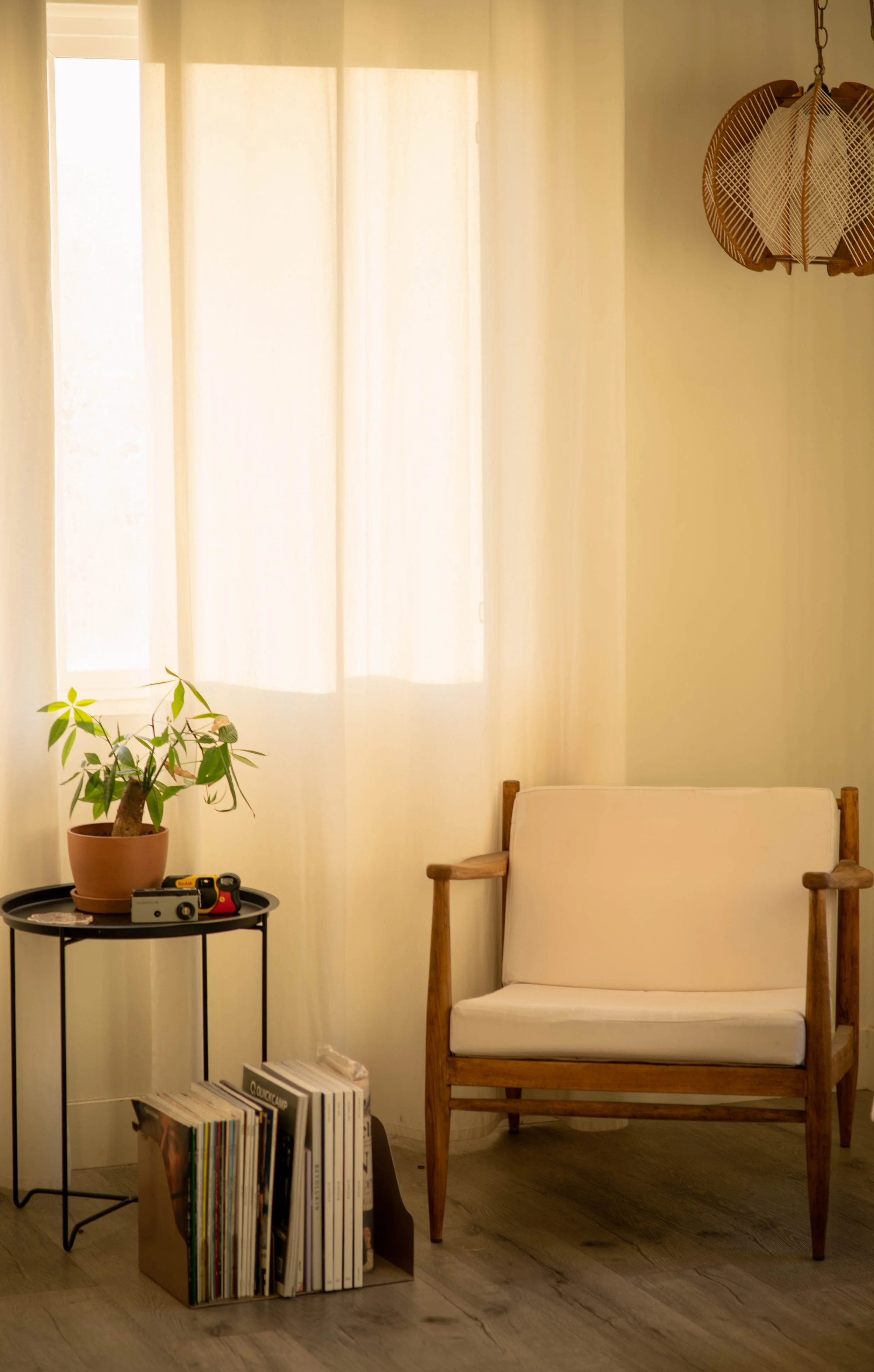A cozy corner of a room with a window covered by light curtains, a wooden armchair with white cushions, a small black round side table holding a potted plant, a vintage camera, and some books or magazines in a paper bag on the floor.