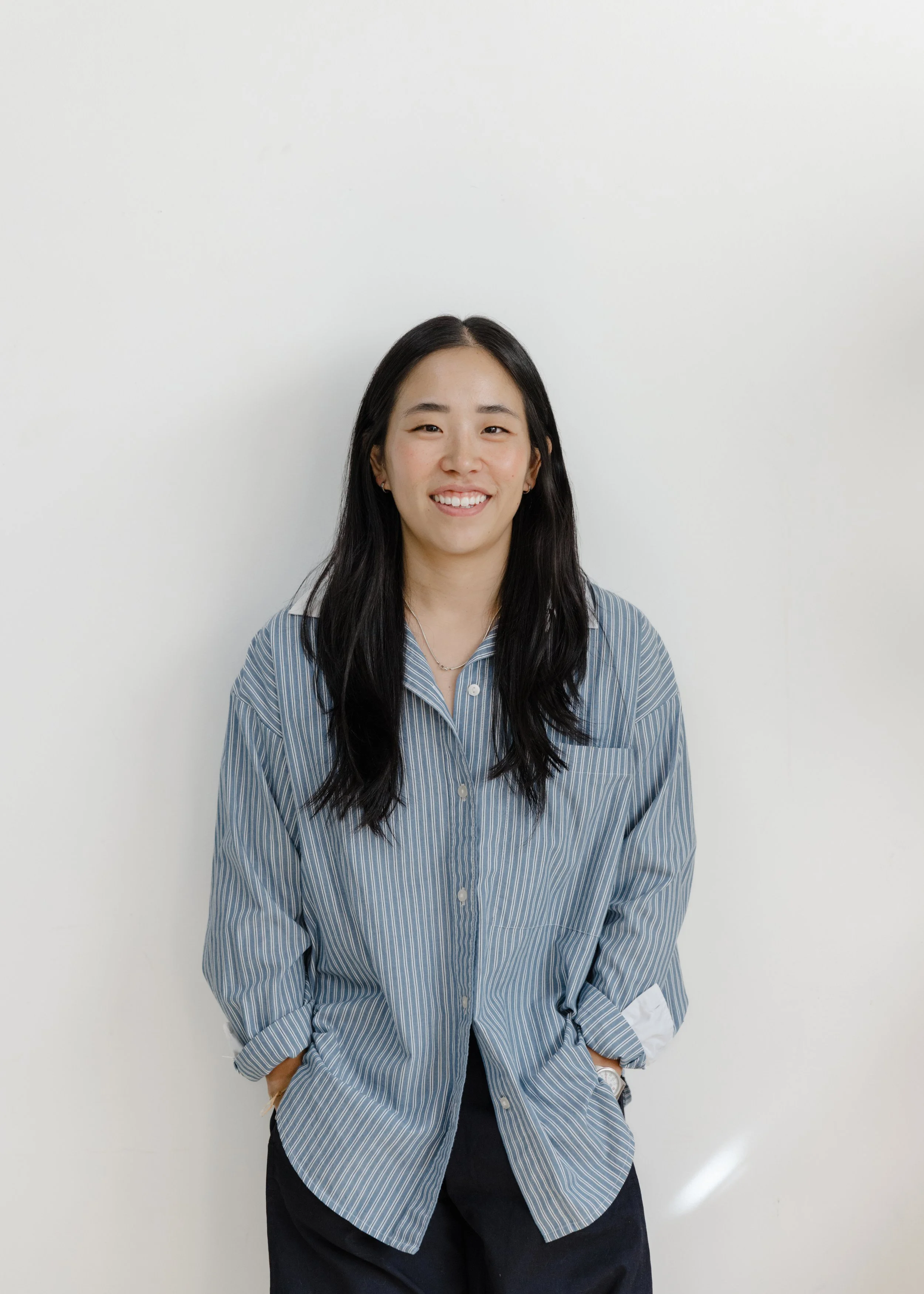 Young woman with long black hair smiling, wearing a blue and white striped button-up shirt and dark pants, standing against a plain white wall.