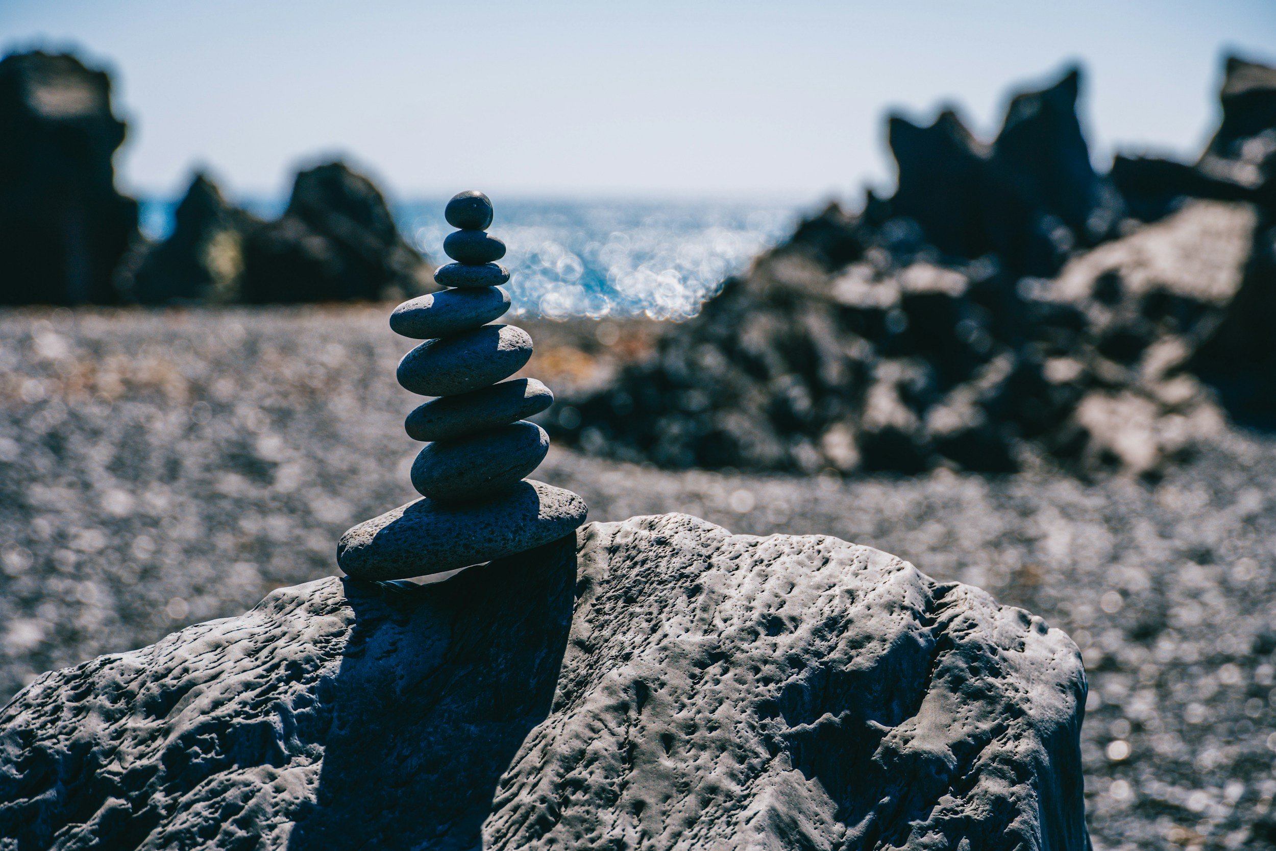 Stacked black stones balance on a large white rock at a rocky beach with the ocean and distant rocks in background.