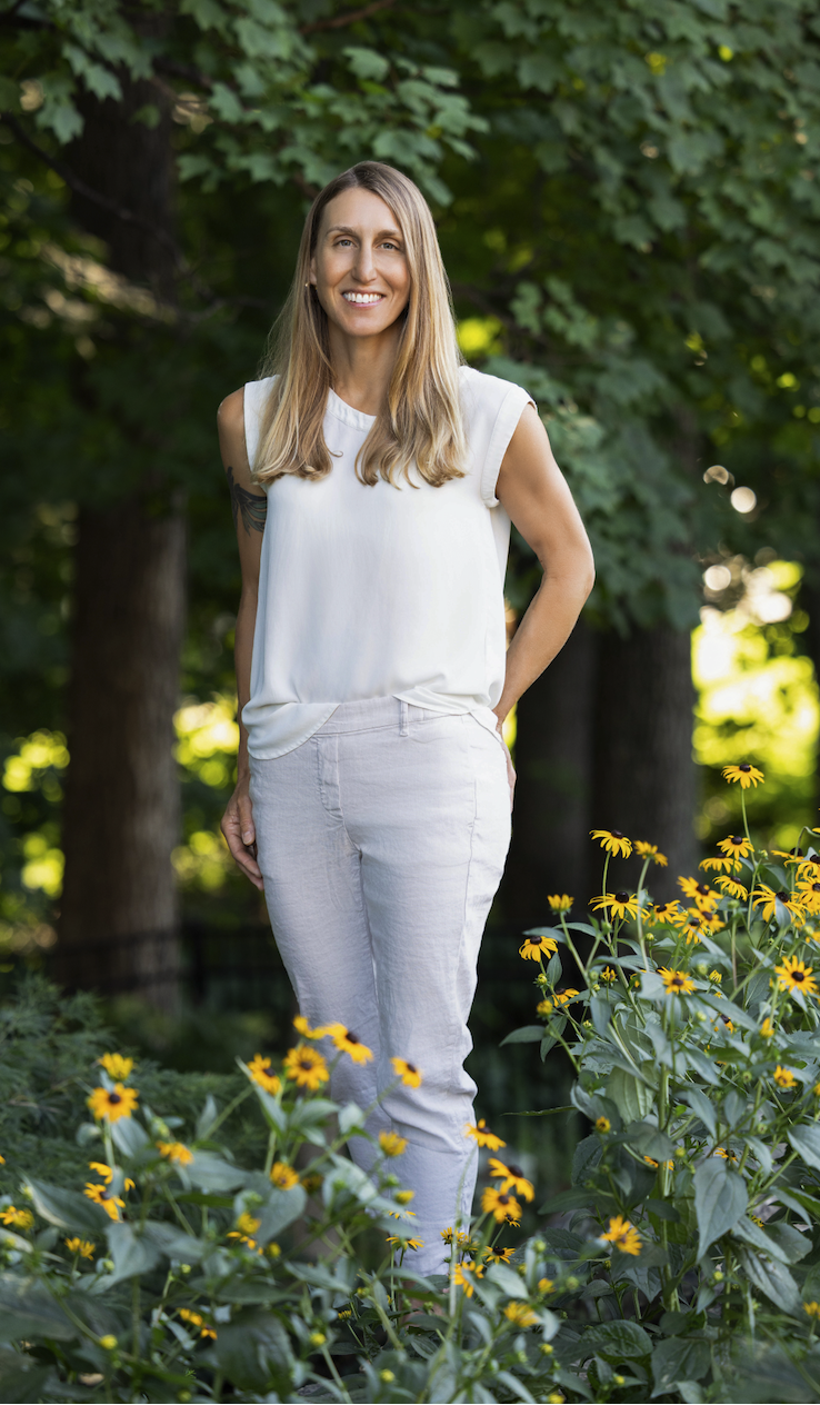Woman standing outdoors among yellow flowers and green foliage in a park or garden during daytime.