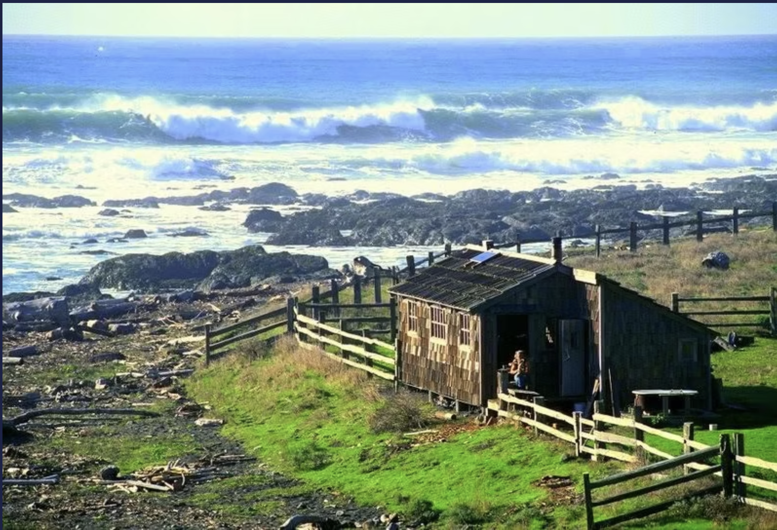 A small wooden house on a grassy hill near the shoreline with rocks and driftwood, overlooking ocean waves under a partly cloudy sky.