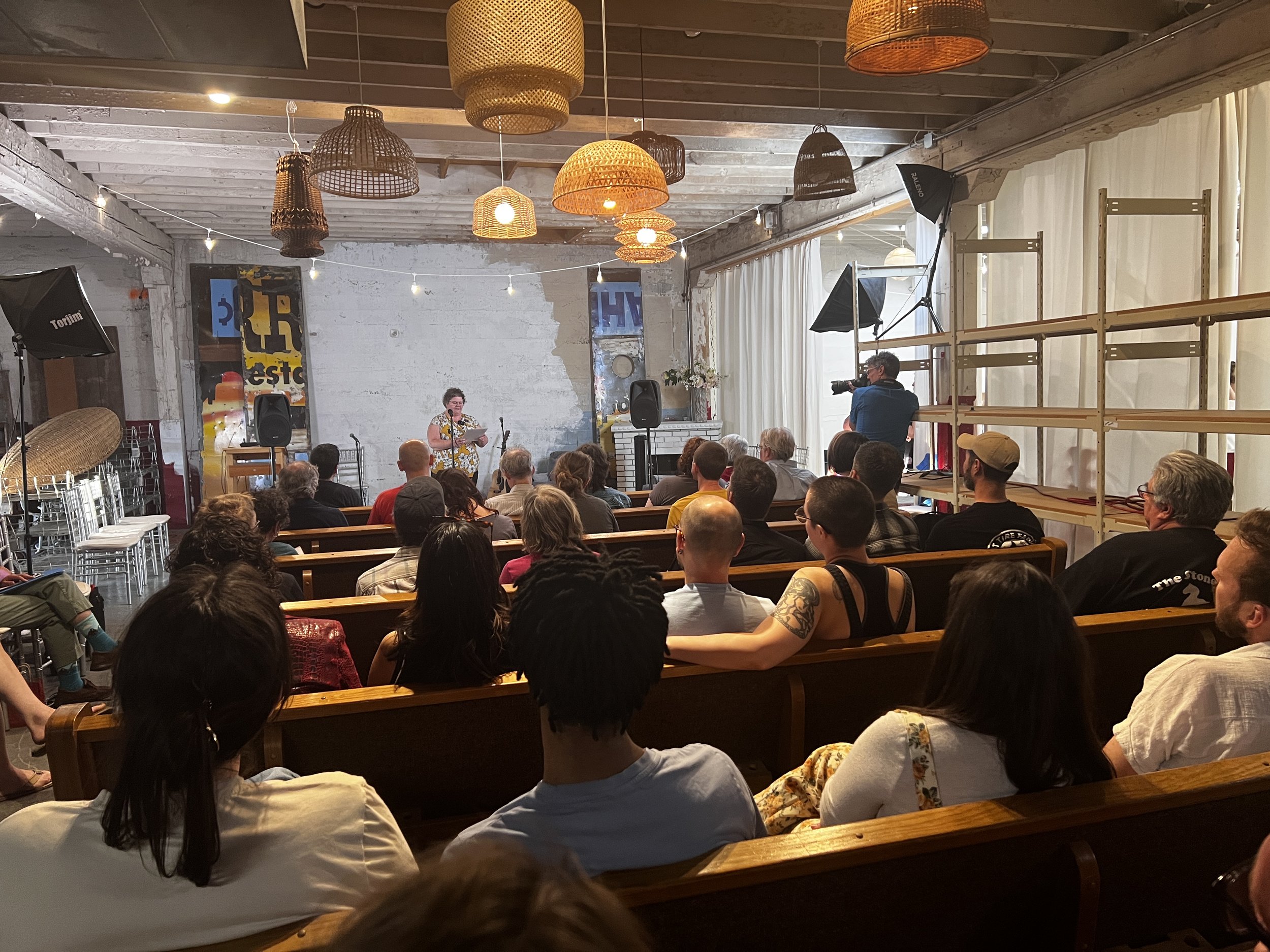 Audience attending a poetic reading or performance in a rustic, cozy space with hanging woven lights and textured white brick walls.