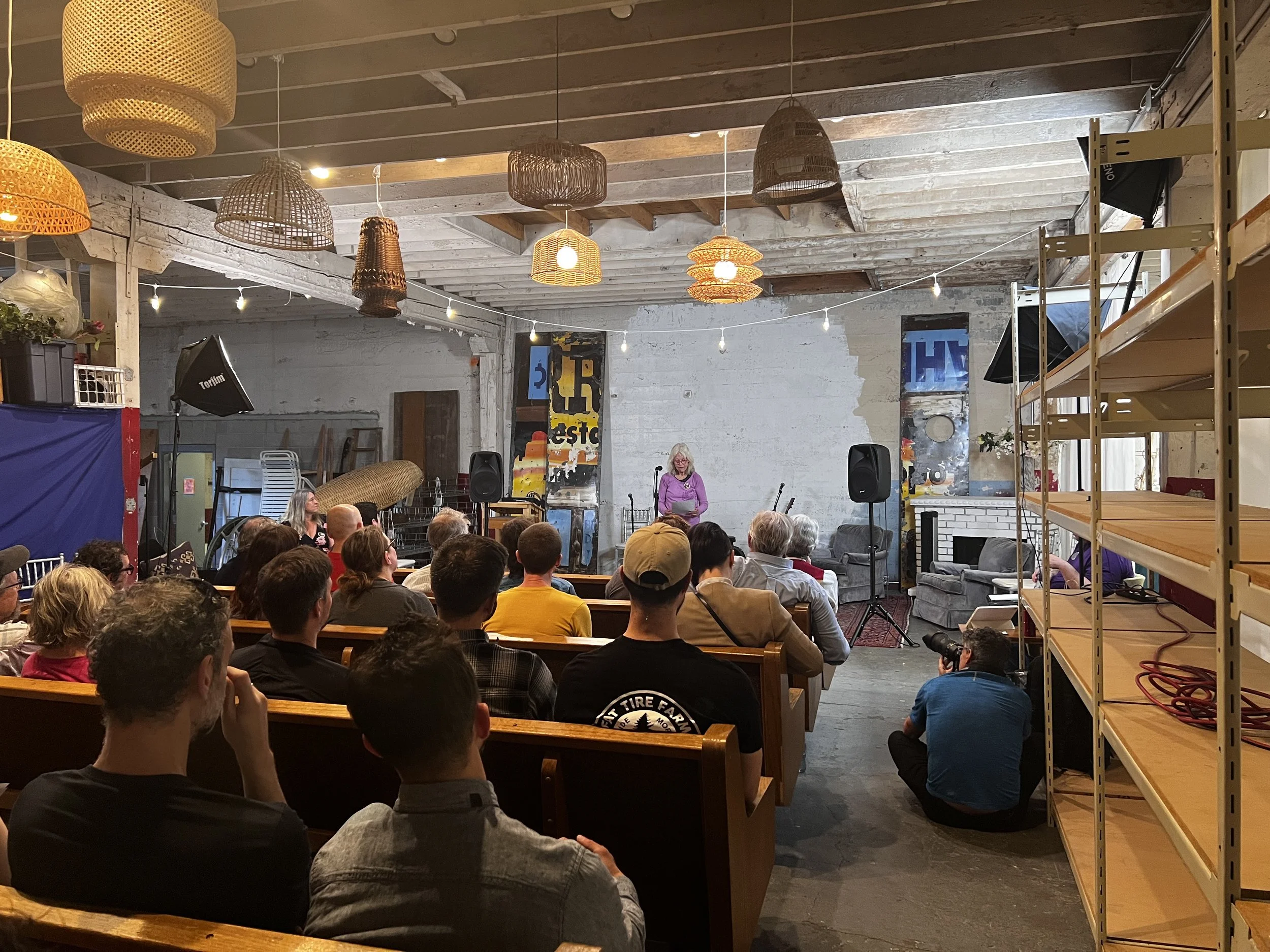 An indoor gathering with an audience seated on benches, facing a woman standing at a microphone in front of a white brick wall in a decorated room with hanging woven lamps.