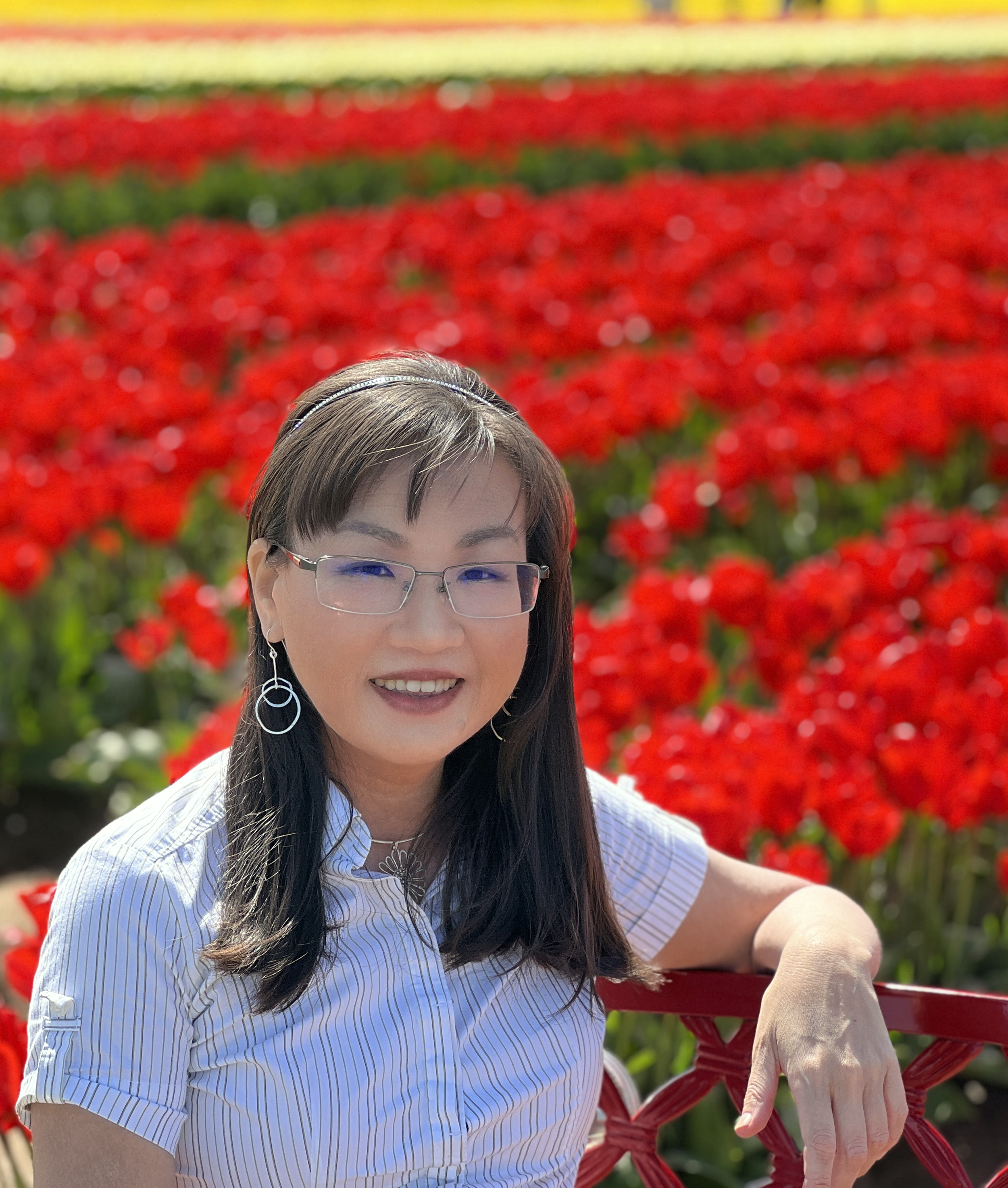 A woman with glasses and earrings smiling outdoors sitting on a red bench in a tulip garden with red tulips in the background.