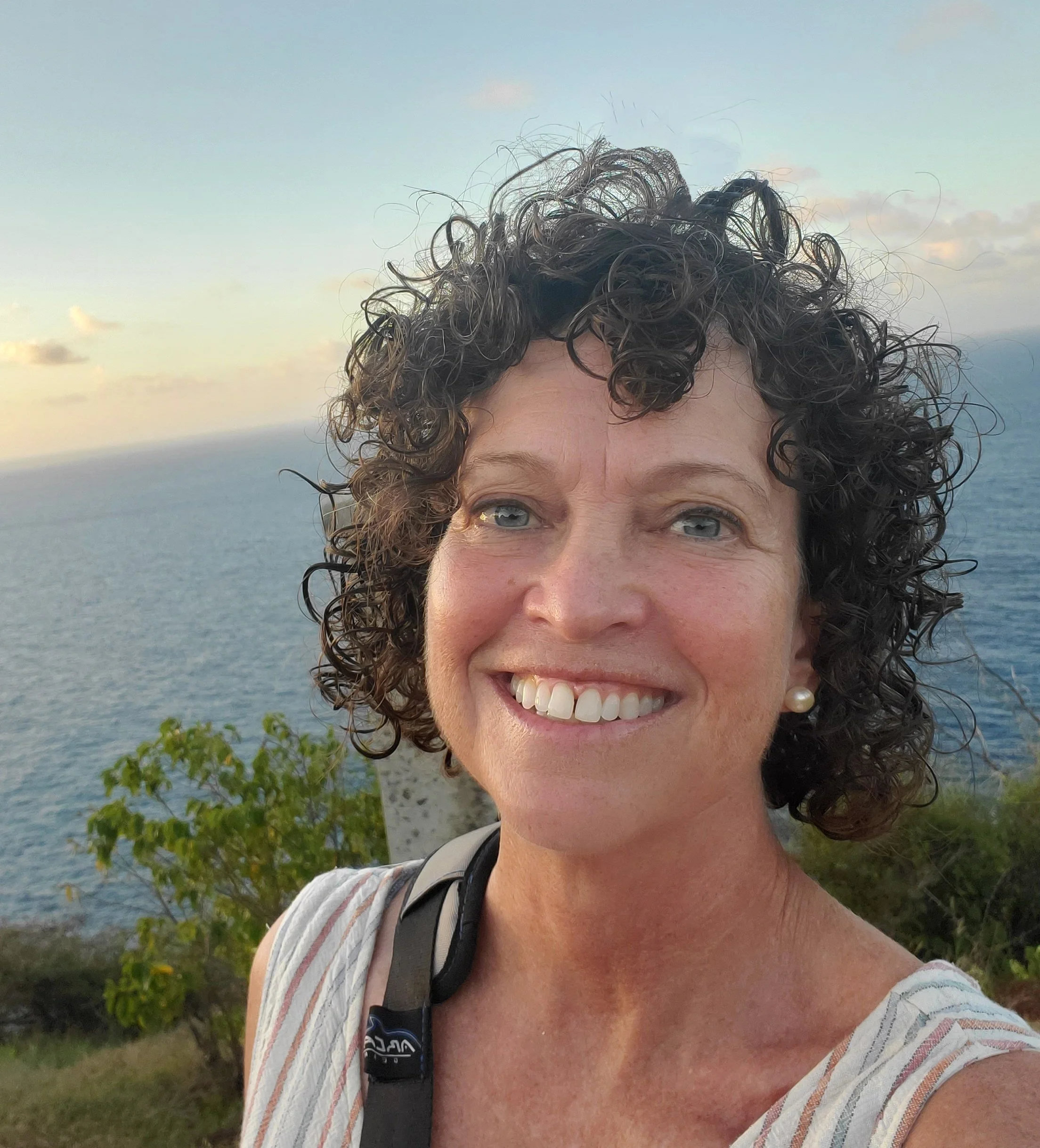 A woman with short, curly dark hair and blue eyes taking a selfie outdoors near the ocean, with a blue sky and some clouds in the background.