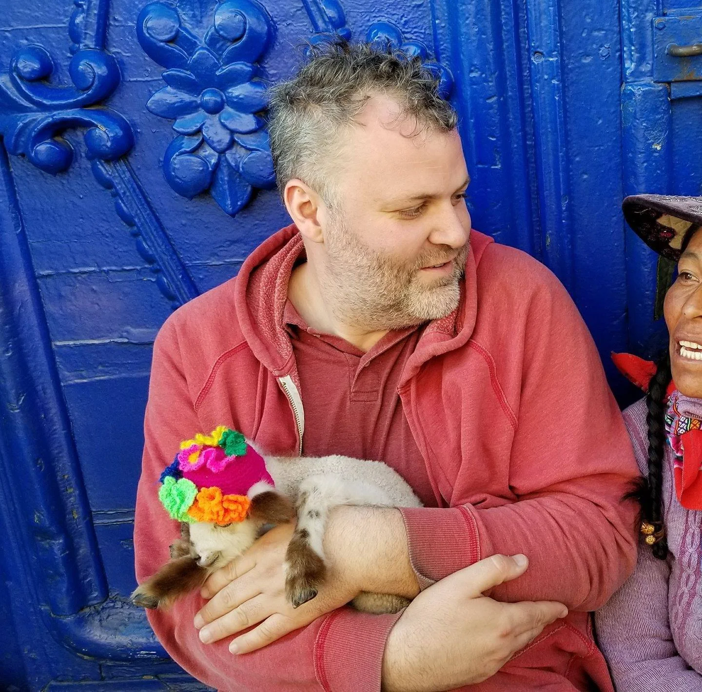 A man with gray hair and a beard holding a small rabbit, wearing a colorful knitted hat, in front of a blue decorative wall, with a woman partially visible on the right side of the image.
