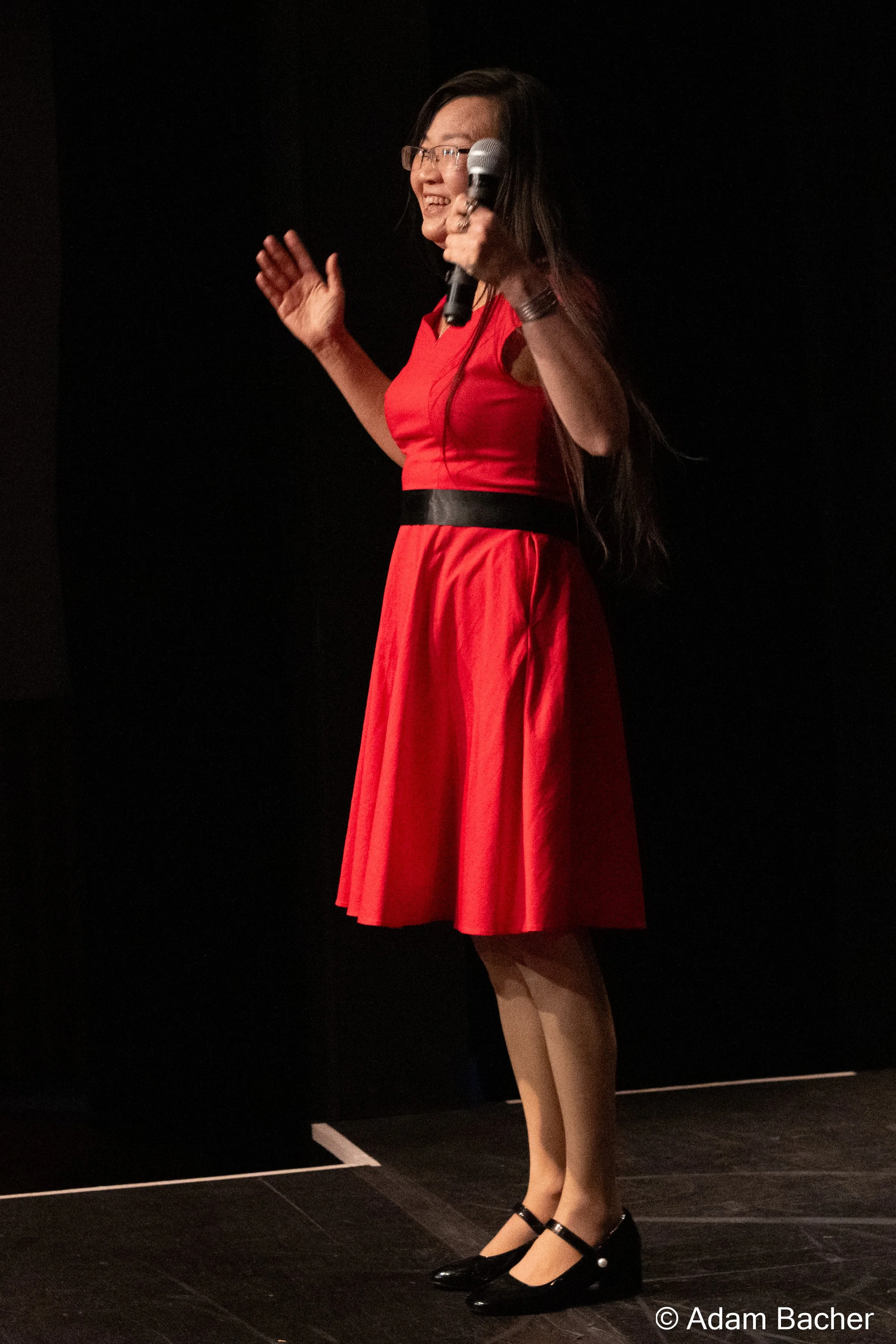 A woman in a red dress holding a microphone on stage, smiling, and waving.