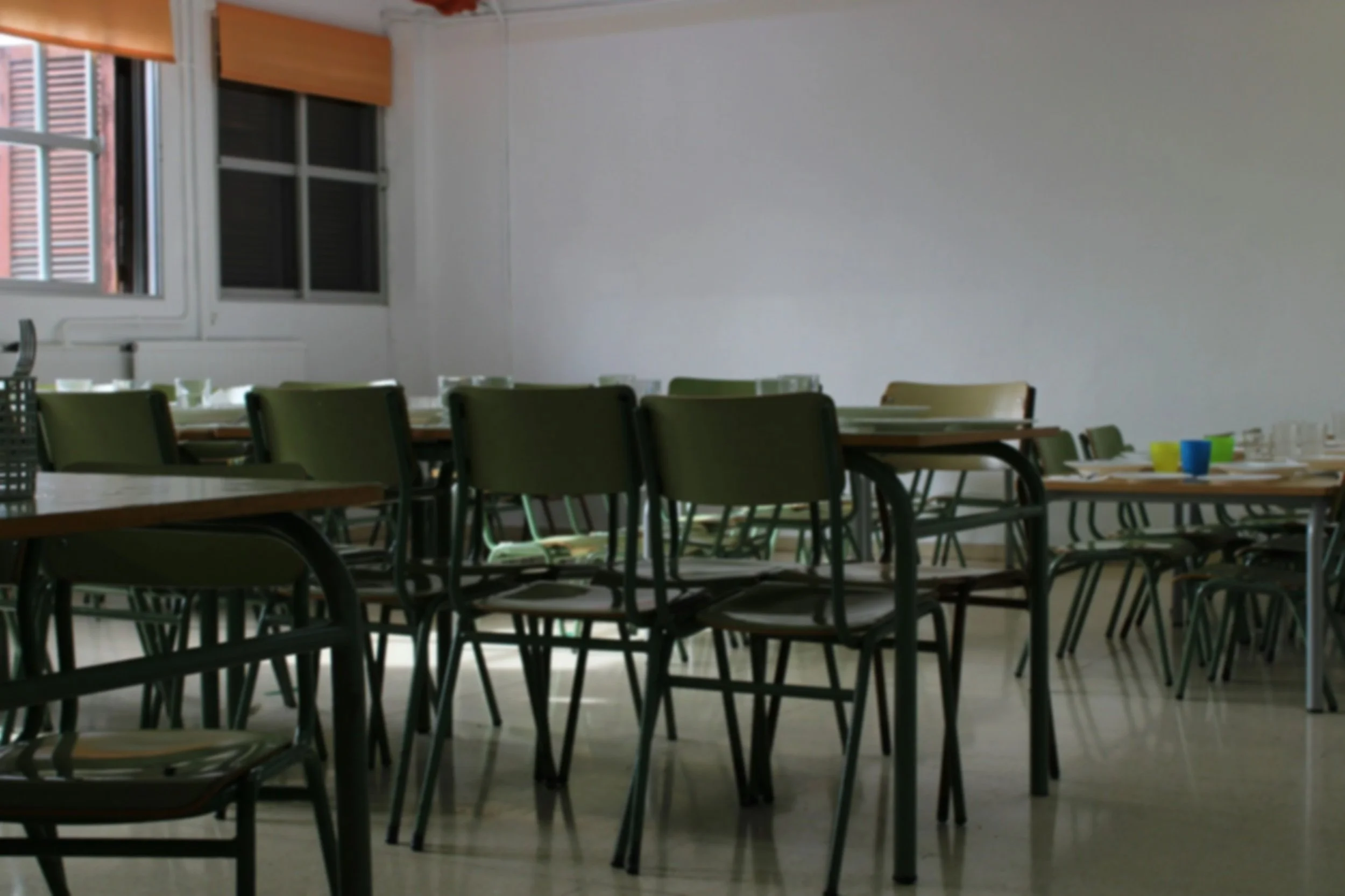 Empty dining room with tables and green chairs, window with blinds, and cups on the tables.