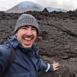 A smiling man with dark hair, glasses, and a beard, wearing a dark jacket, outdoors with a cloudy sky in the background.