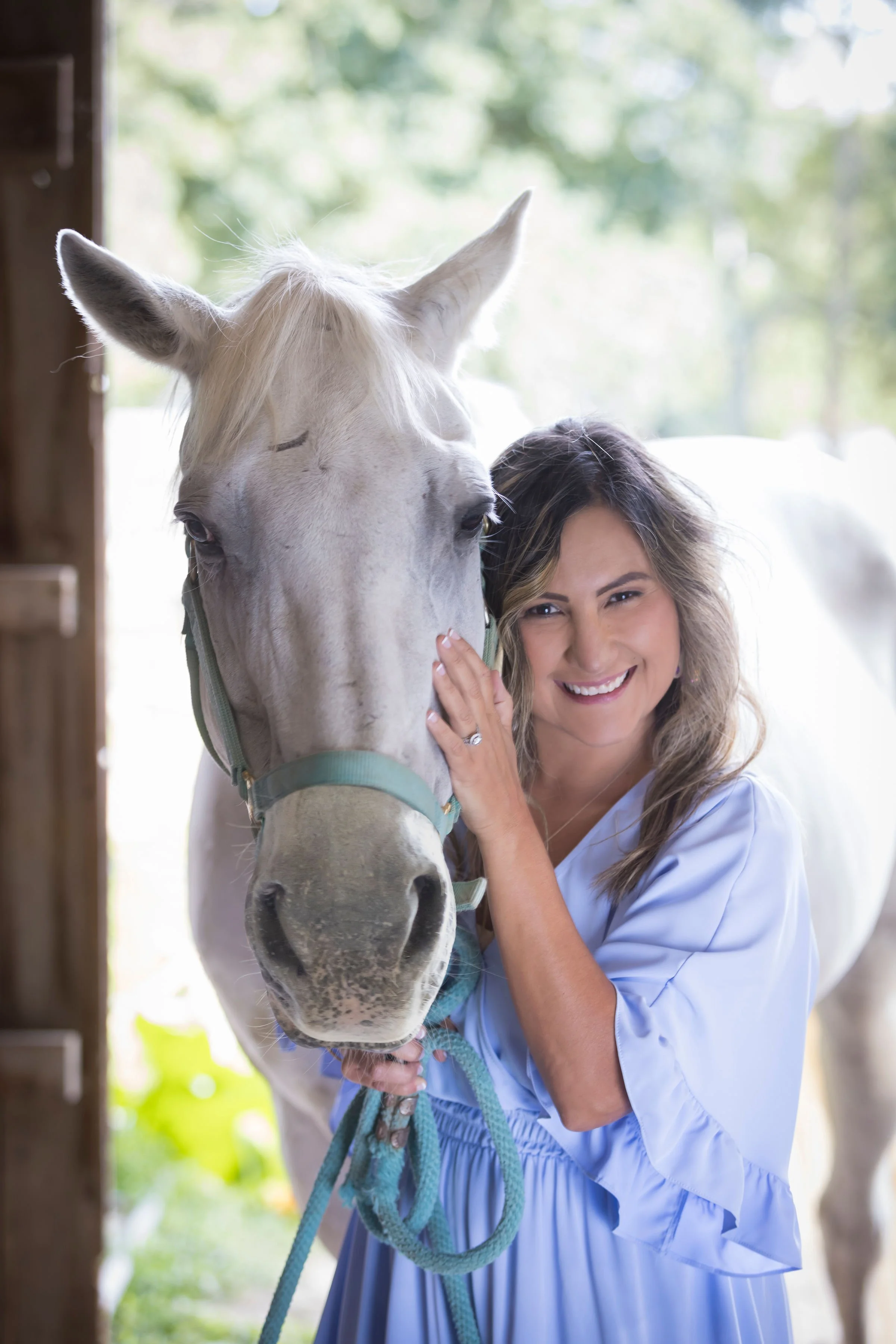 A woman in a light blue dress smiling and winking while holding a white horse's face, standing inside a wooden stable with sunlight filtering through trees outside.