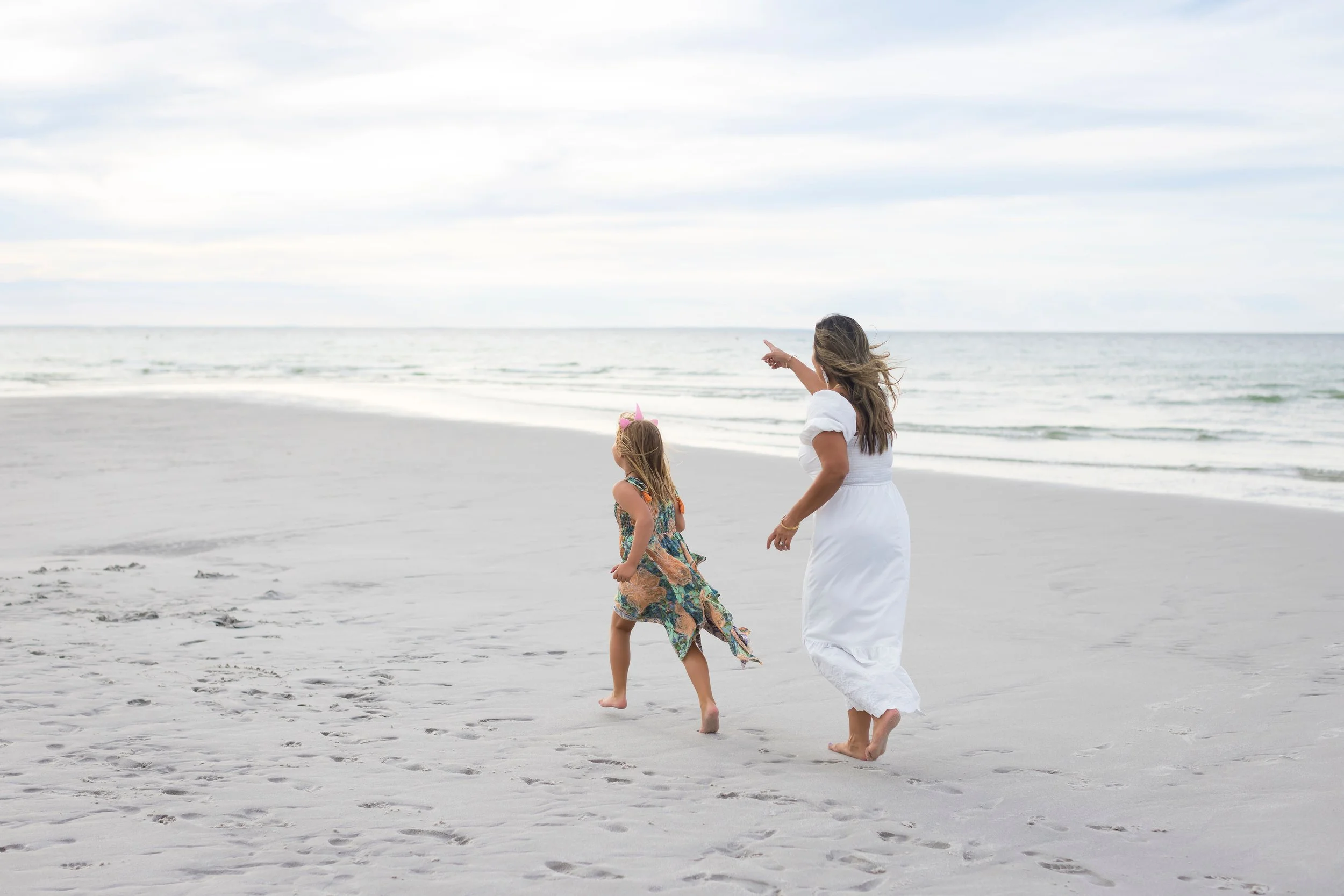 A mother and a young daughter running along the beach, the woman is pointing towards the ocean.