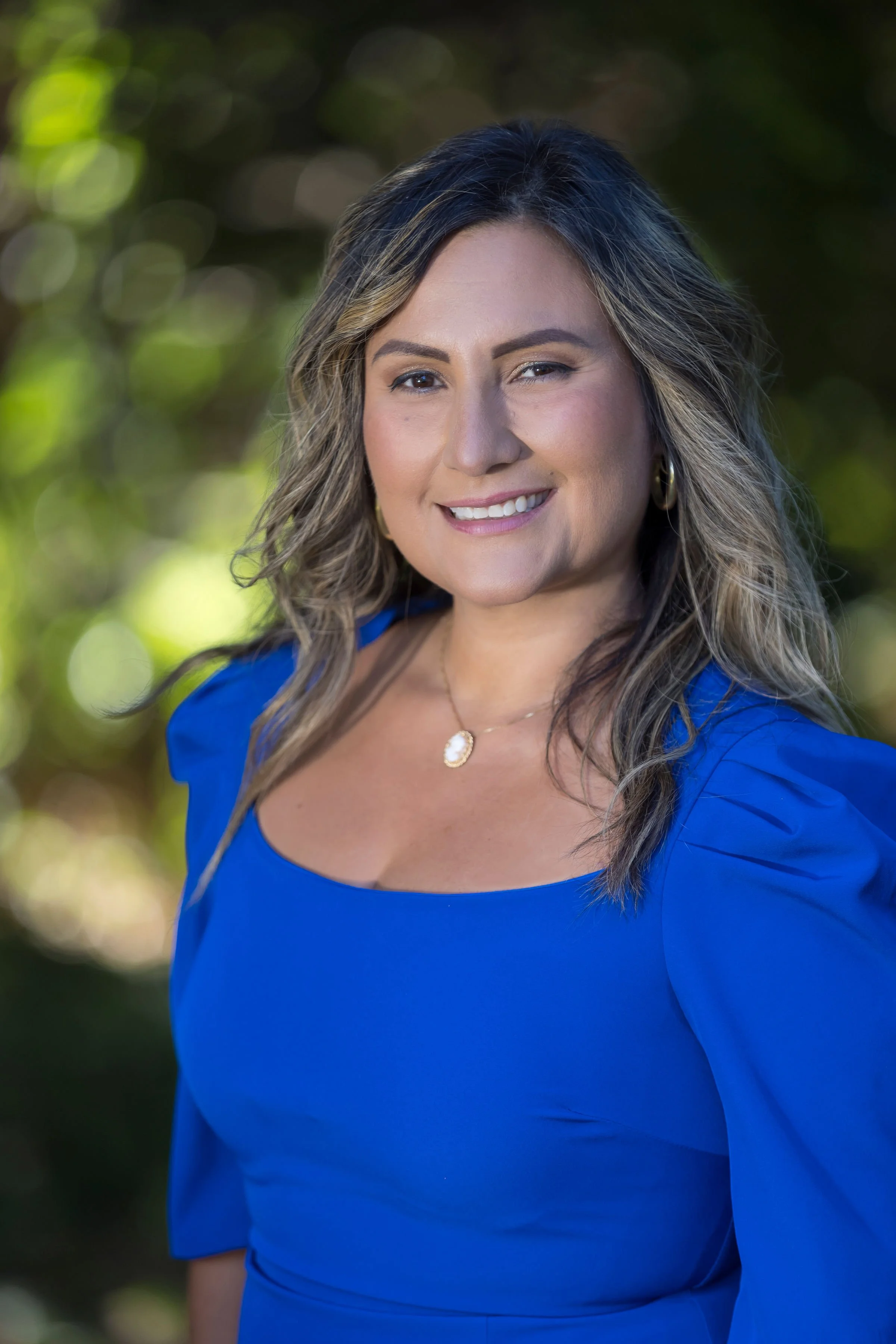 Portrait of a female psychiatrist  in a bright blue dress, smiling, with wavy hair, outdoor background with blurred green foliage.