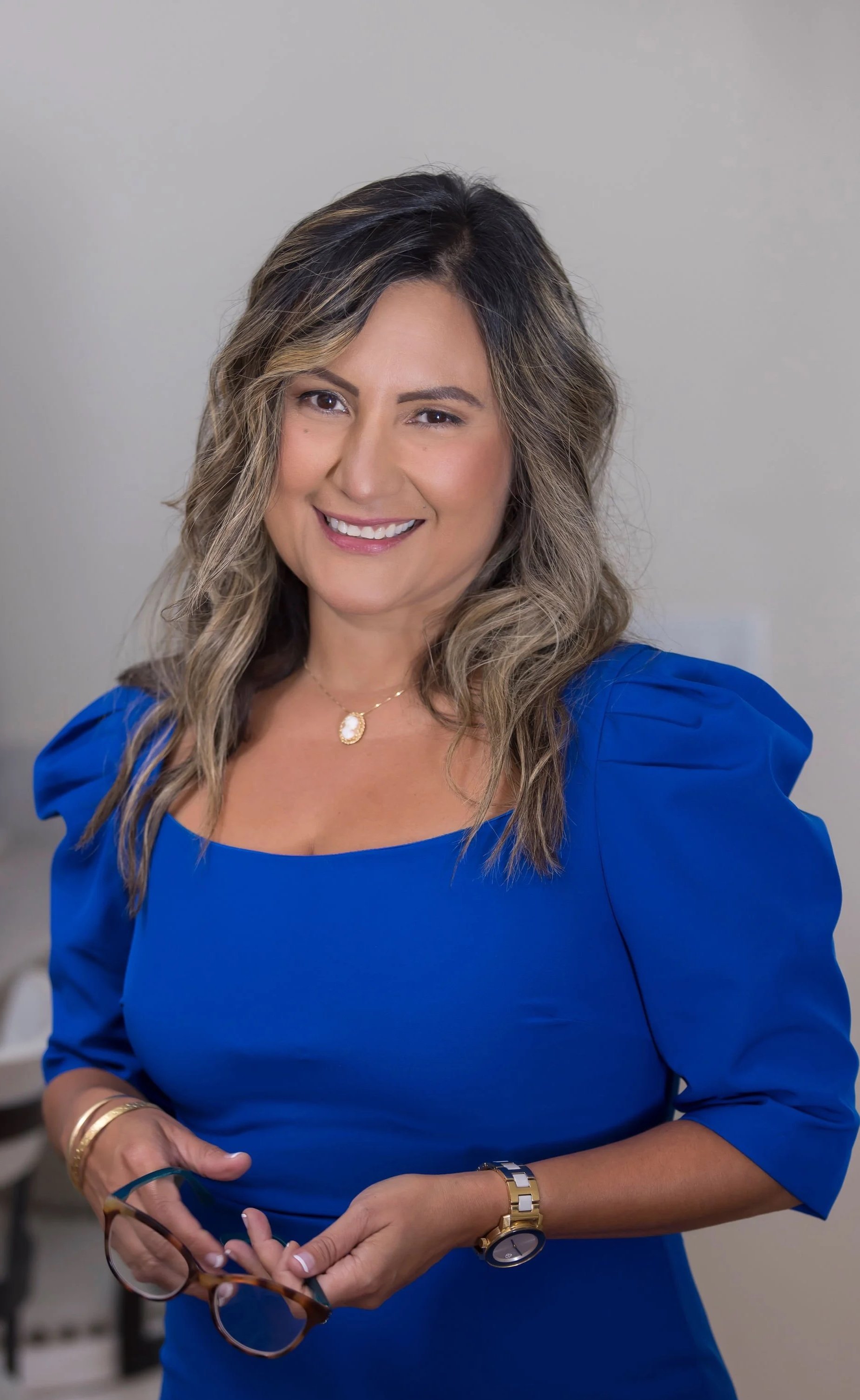 A woman with wavy brown hair wearing a royal blue dress, smiling, holding glasses, with jewelry including a necklace, bracelet, and watch.