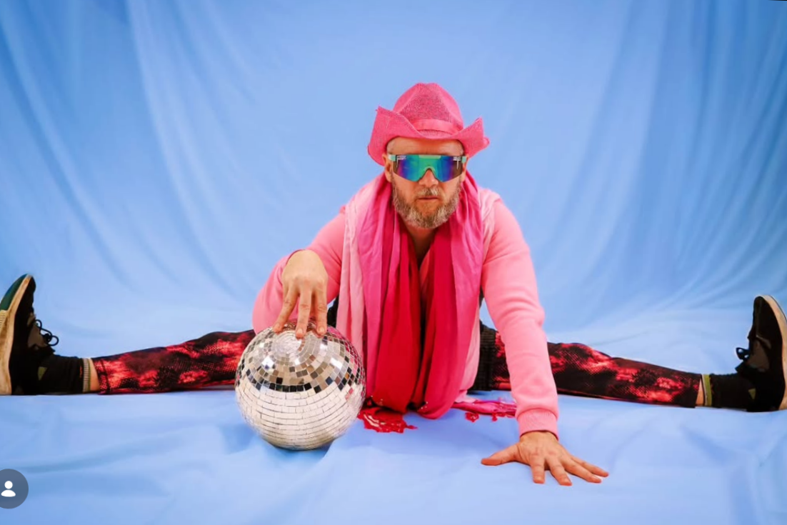 A man in colorful clothing and a pink cowboy hat is doing a split while holding a disco ball, with a blue background behind him.