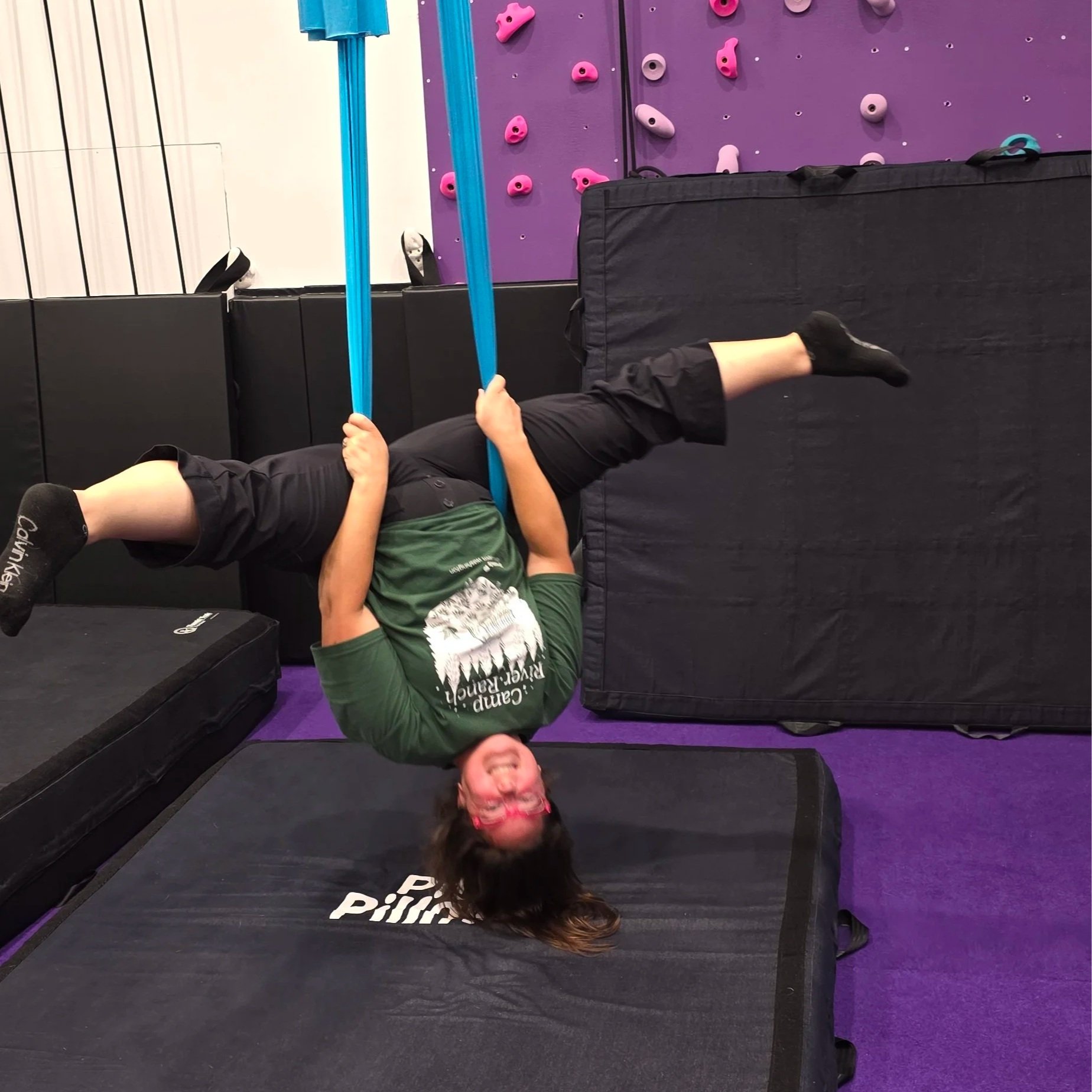 Person lying on the floor underneath a padded black crash mat at an indoor climbing gym, holding onto blue climbing holds on a purple climbing wall, with a smiling face and pink glasses.
