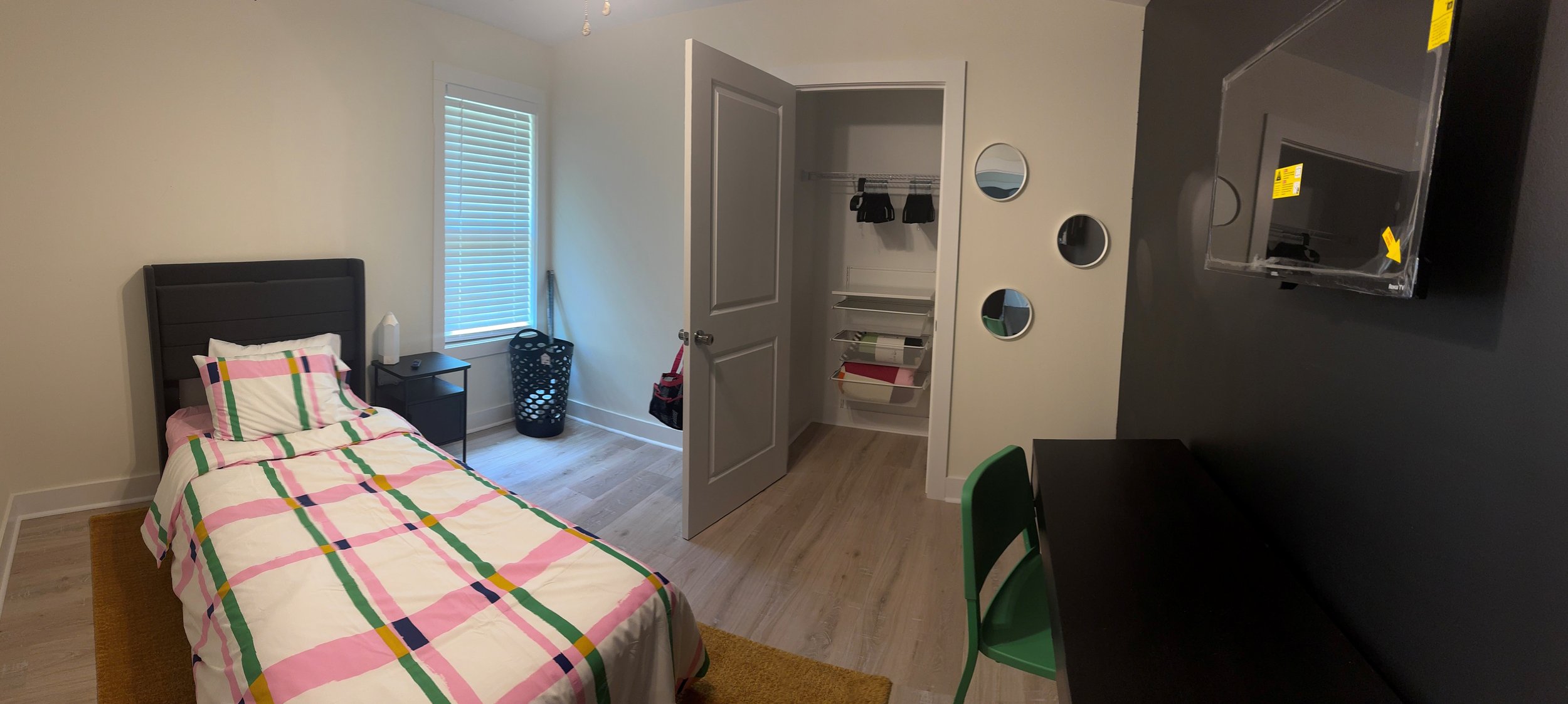 A bedroom with a single bed, a window with blinds, a nightstand with a lamp, an open closet with shelves, three small circular mirrors on the wall, a black dresser with a television, and a green chair.