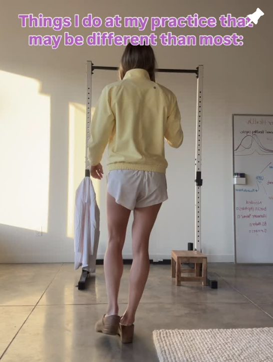 A woman in a yellow jacket and white shorts standing in a gym, facing away from the camera, with a whiteboard and workout equipment in the background.