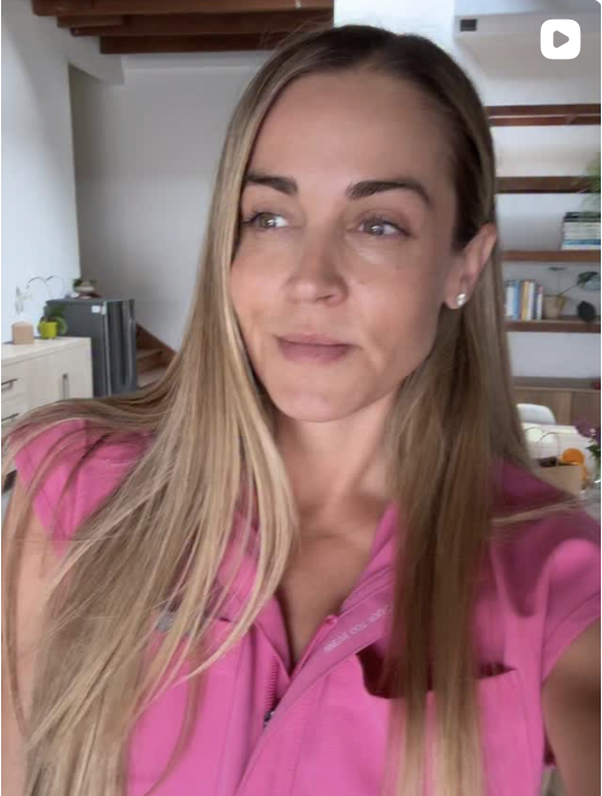 A woman with long blonde hair and earrings wearing a pink shirt taking a selfie indoors with a background of wooden shelves and books.