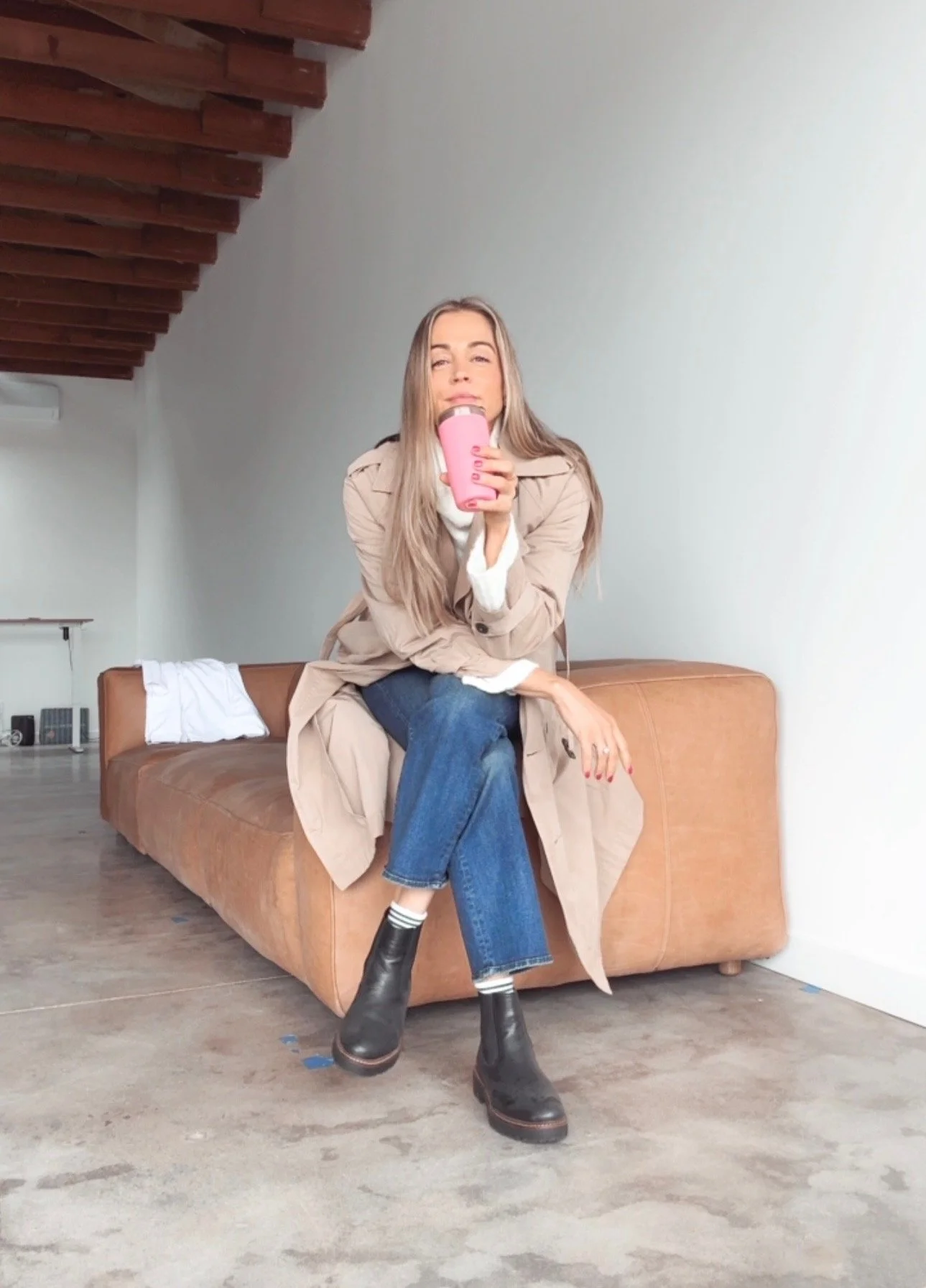 A woman with long blonde hair sitting on a brown leather couch, holding a pink travel mug, wearing a beige trench coat, blue jeans, and black ankle boots, in a room with a white wall and wooden ceiling beams.