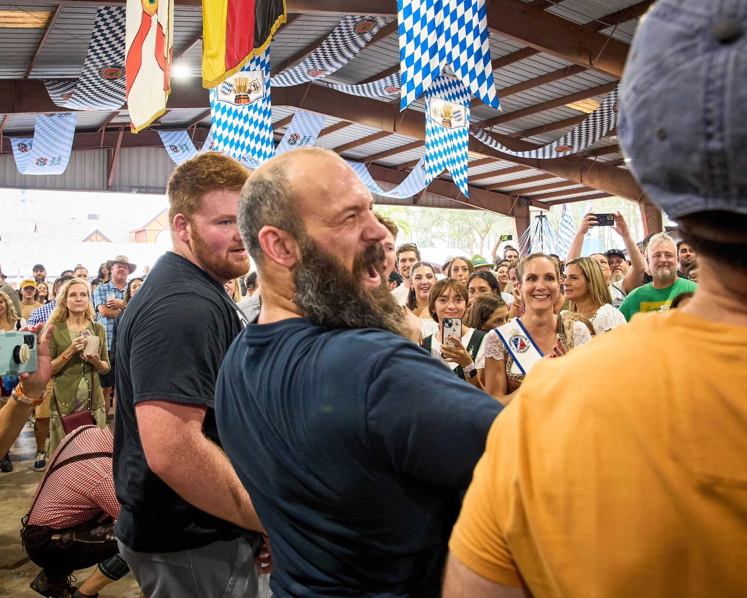 Crowd of people at an indoor event decorated with blue and white checkered banners and flags, many smiling and taking photos, with some wearing traditional Bavarian clothing.