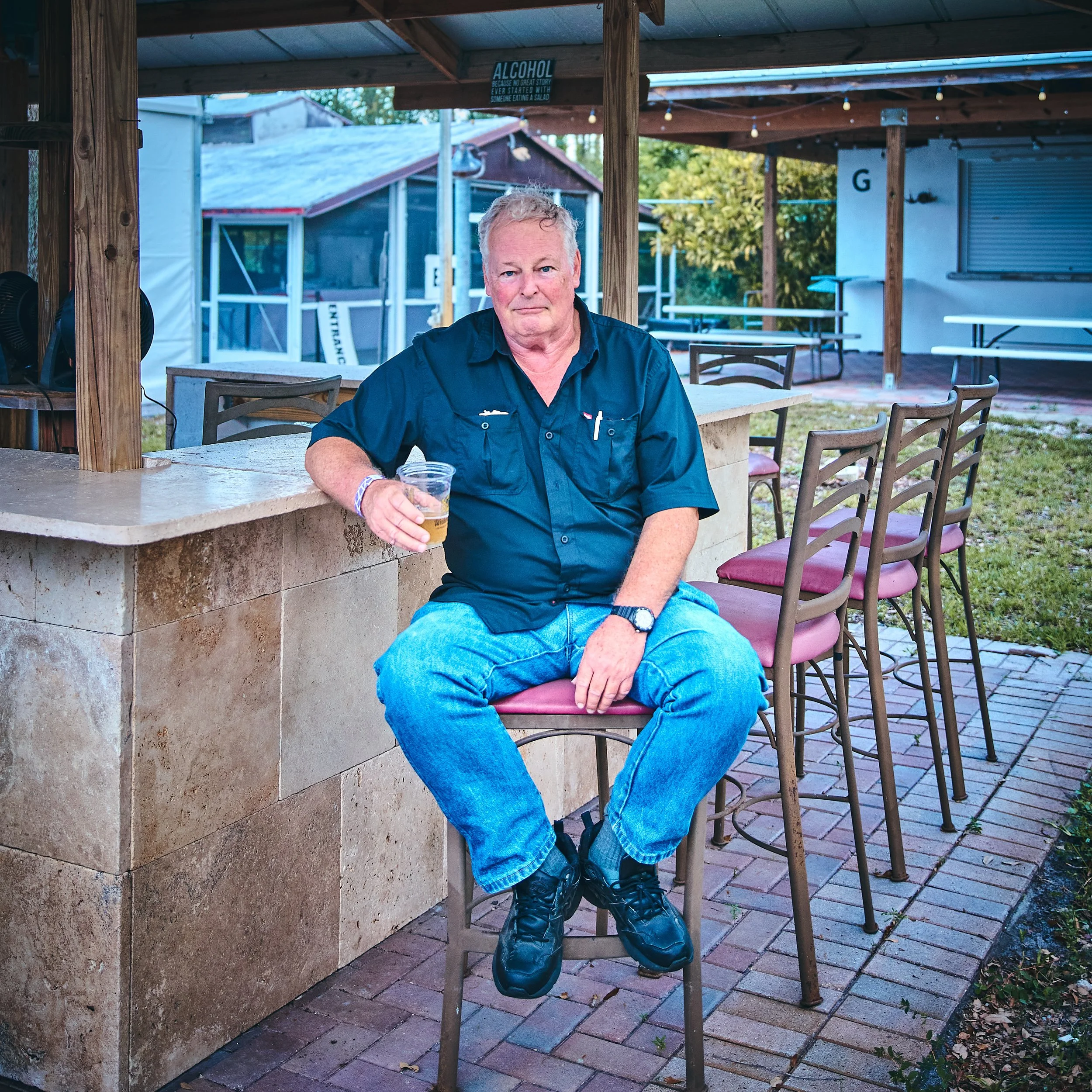 A man with gray hair sits on a barstool outdoors at a bar or restaurant, holding a plastic cup with a drink. He is wearing a dark blue shirt, jeans, and black shoes. There are several barstools with pink cushions next to him, and a stone counter behi
