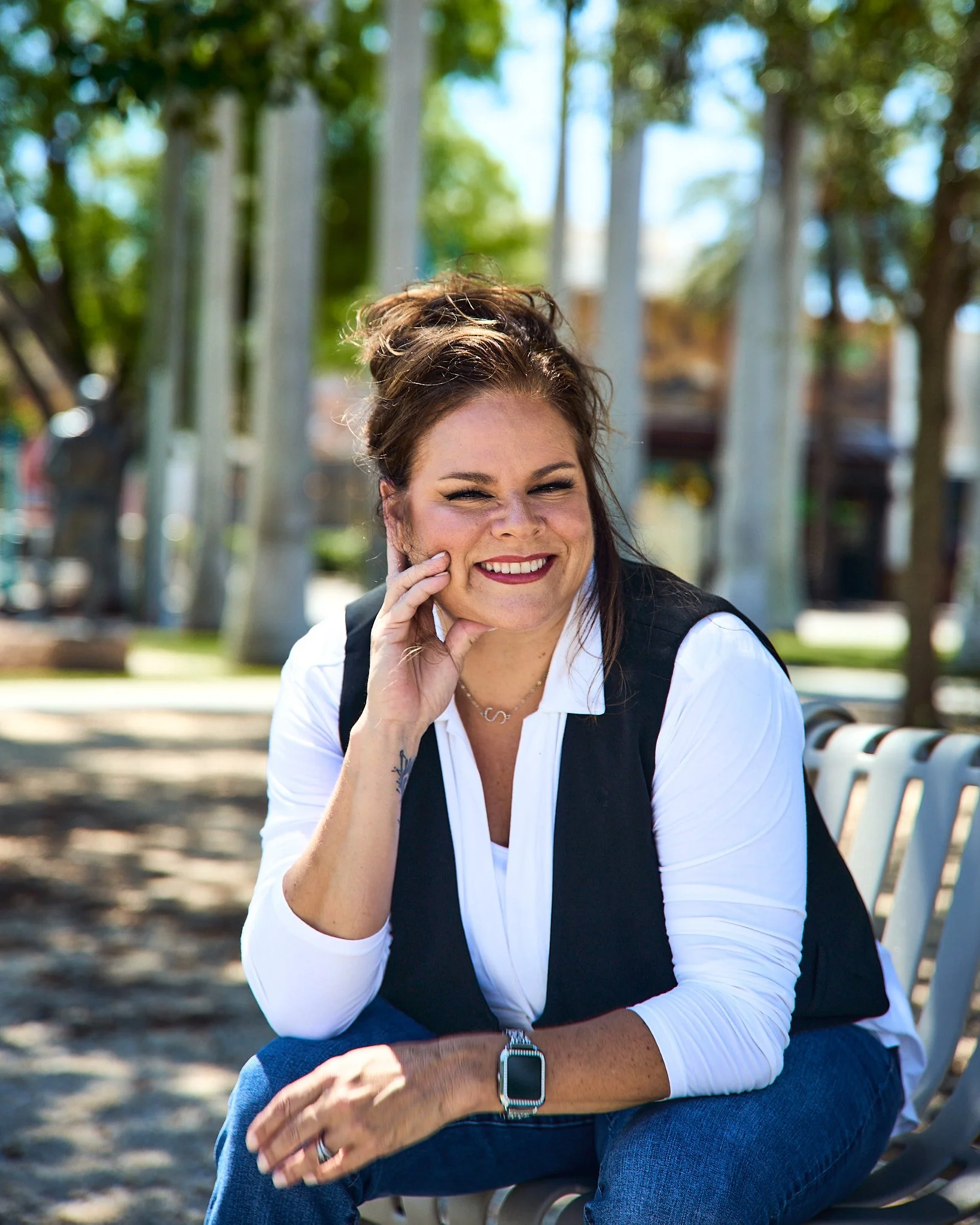 A smiling woman with brown hair styled in a messy bun, wearing a white long-sleeve top under a black vest, sitting on a park bench with trees and a playground in the background.