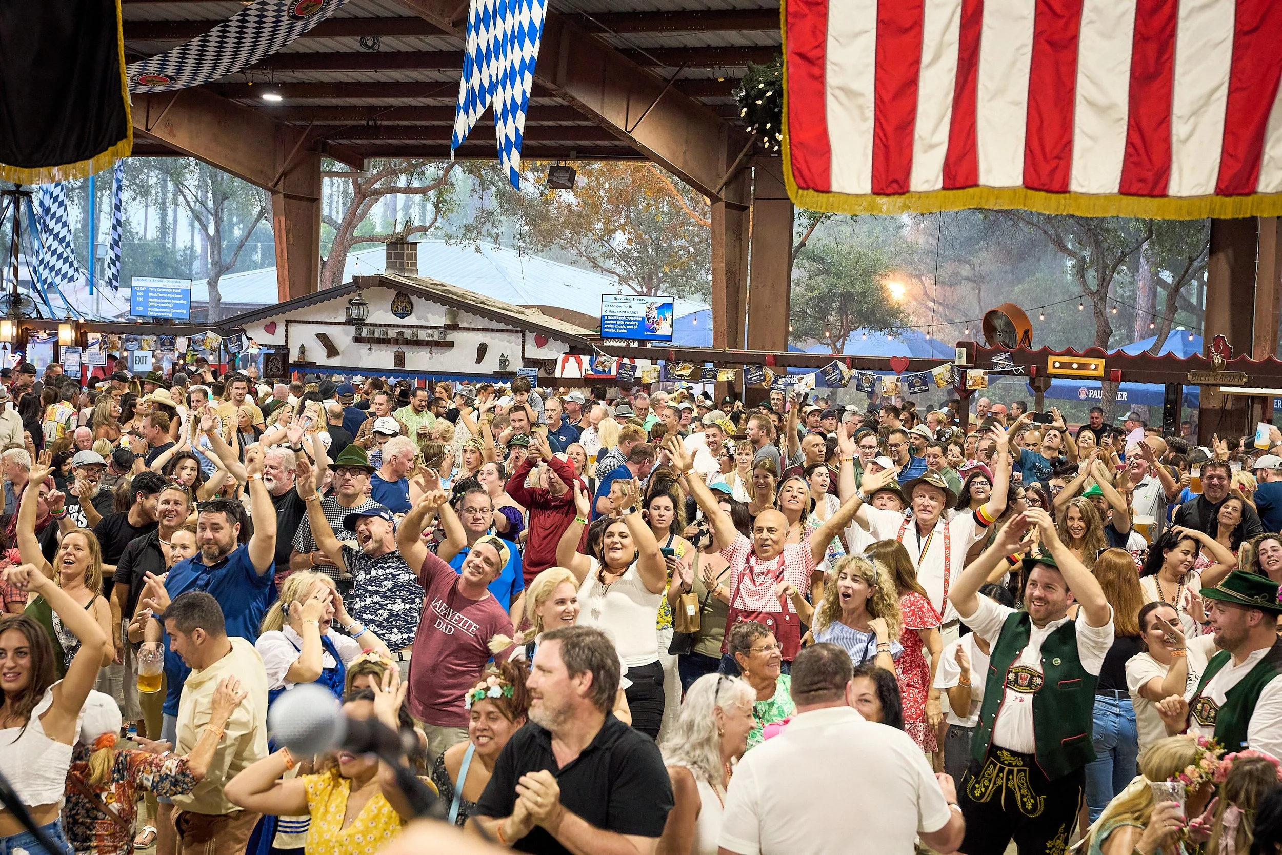 A large crowd of people celebrating at an Oktoberfest event indoors, with traditional German decorations, flags, and attire, including men in lederhosen and women in dirndls.