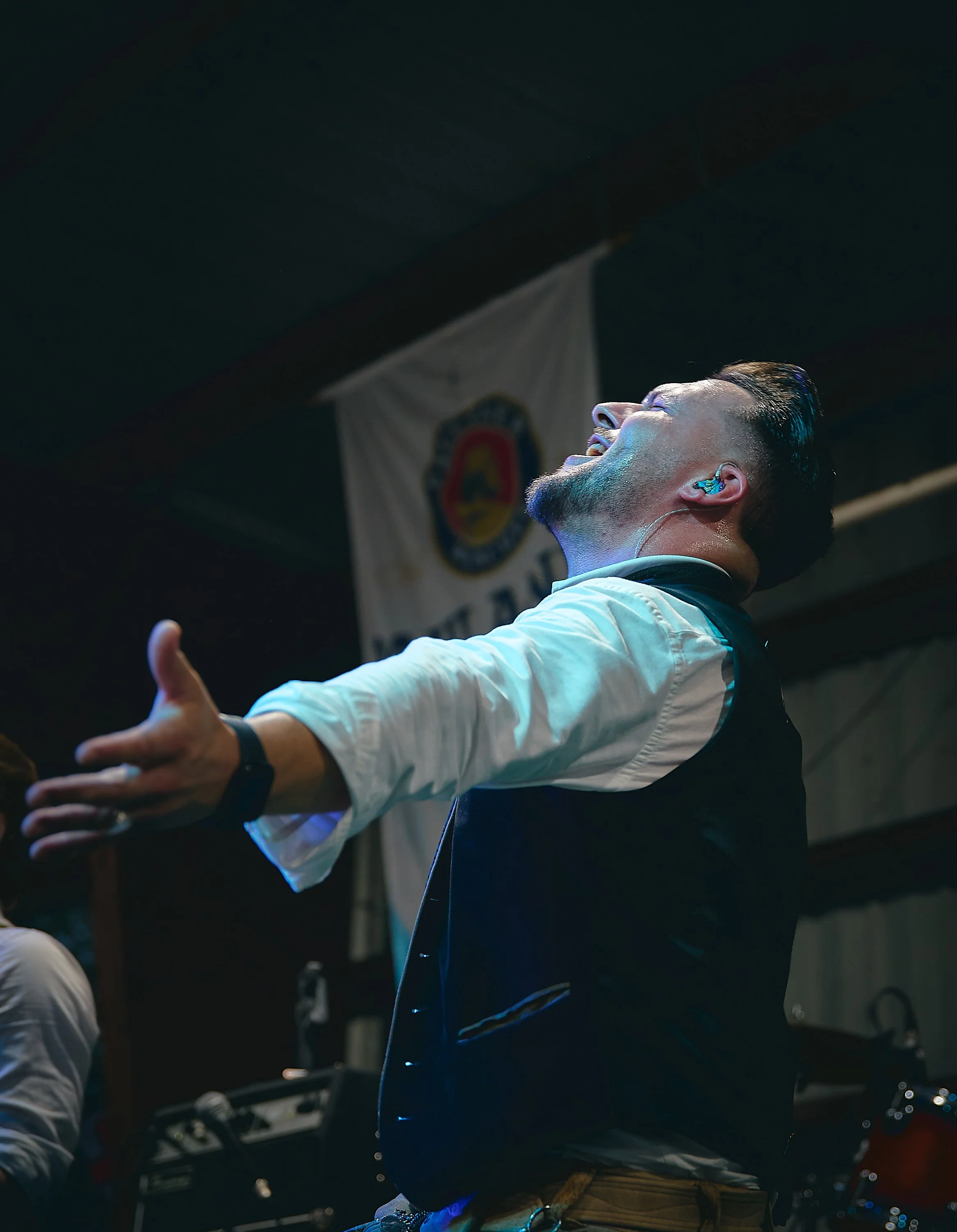 A man on stage, performing passionately with arms outstretched, wearing a white shirt and black vest, with a flag/banner in the background.