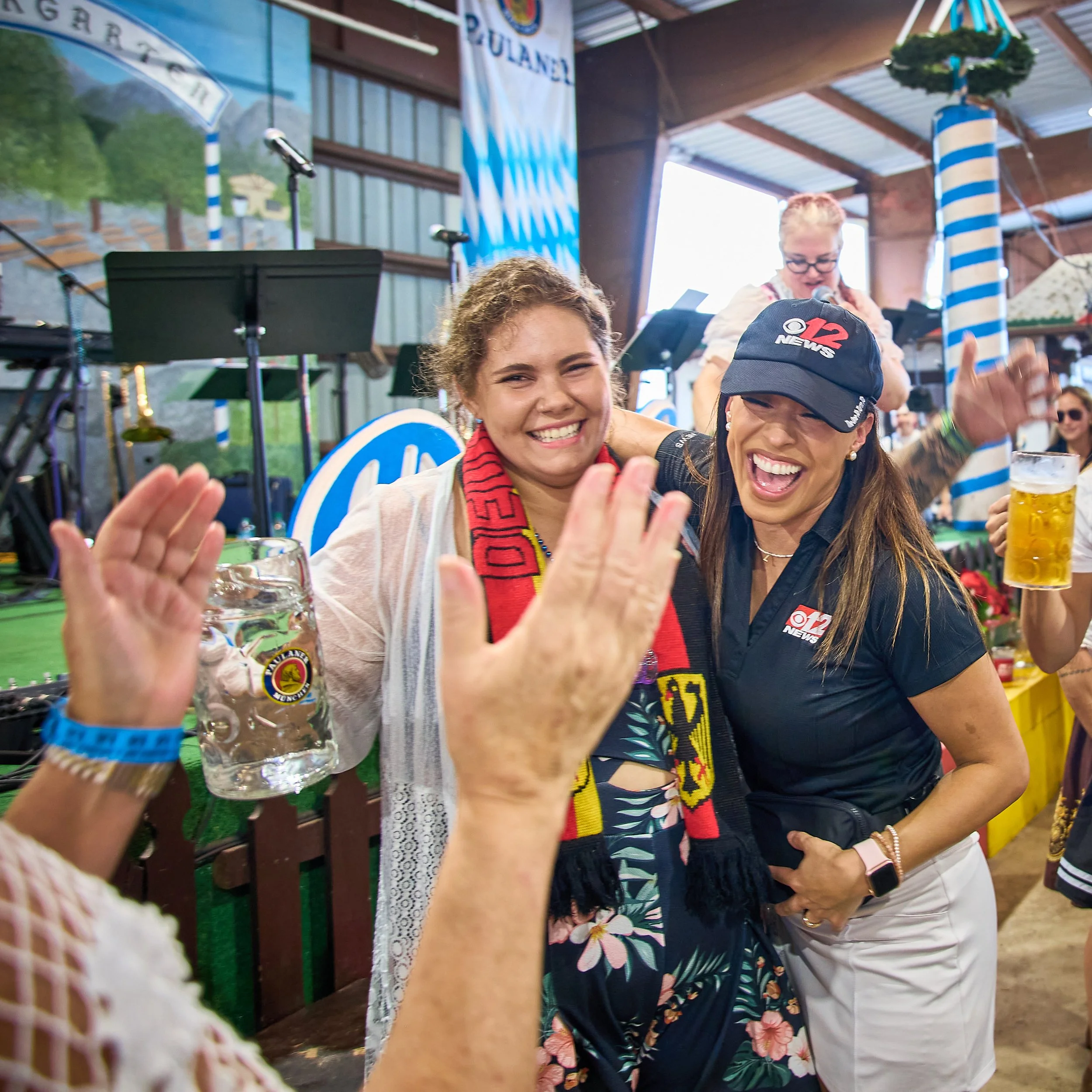 Two women celebrating at a festival, smiling and holding drinks, with a stage and musicians in the background.