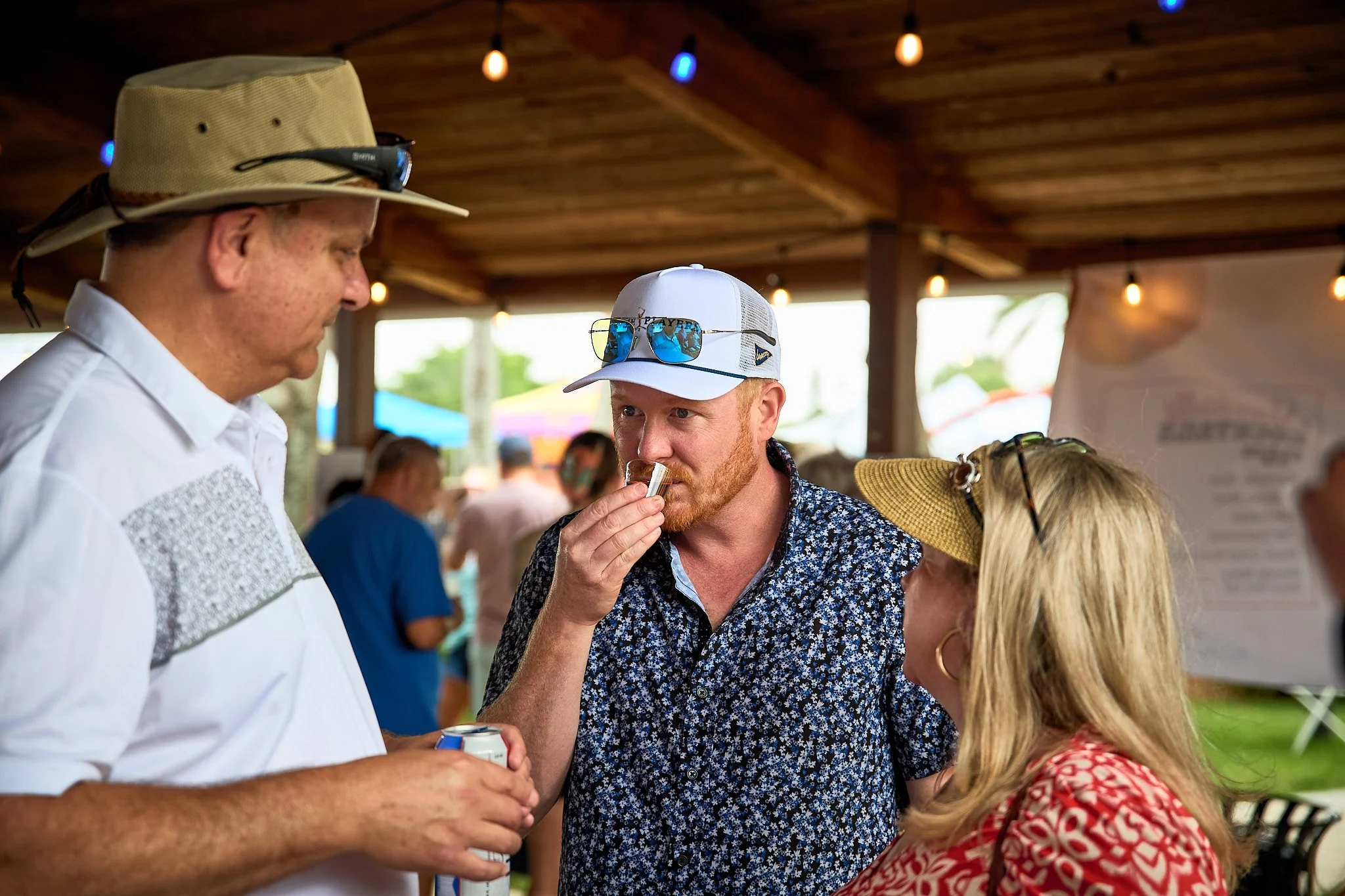 Three people at an outdoor event under a wooden pavilion, two men and a woman, engaged in a conversation, with one holding a beverage can and one tasting food.
